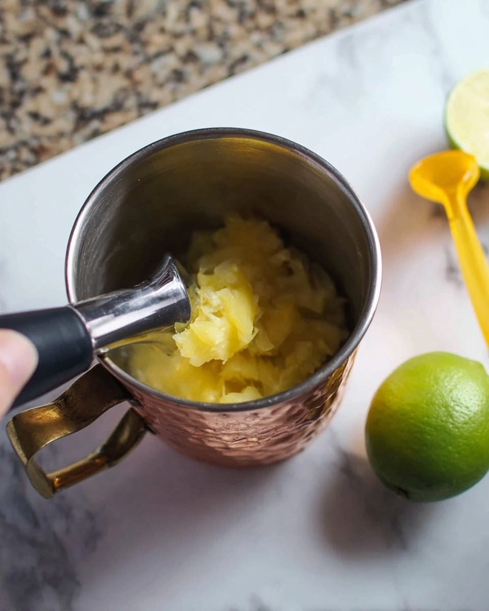 The image shows a metal cup with a copper-colored bottom and a shiny handle on the left side, containing mashed yellow pineapple pieces at the bottom. A woman's hand holds a silver muddler pressing down on the pineapple. To the right on a white marbled surface, there is a whole green lime and a yellow muddler placed sideways. The lighting is natural, showing clear details and texture of the fruit and tools. photo taken with an iphone --ar 4:5 --v 7