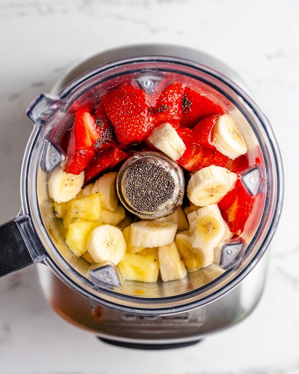 Top view of a metallic blender filled with fruit layers on a white marbled surface. The bottom layer shows a mix of bright red sliced strawberries with black chia seeds sprinkled on top. Above the strawberries are light yellow banana pieces arranged in small chunks. The metal inside of the blender has a shiny, reflective texture. The blender lid is off and set to the side. Photo taken with an iphone --ar 4:5 --v 7
