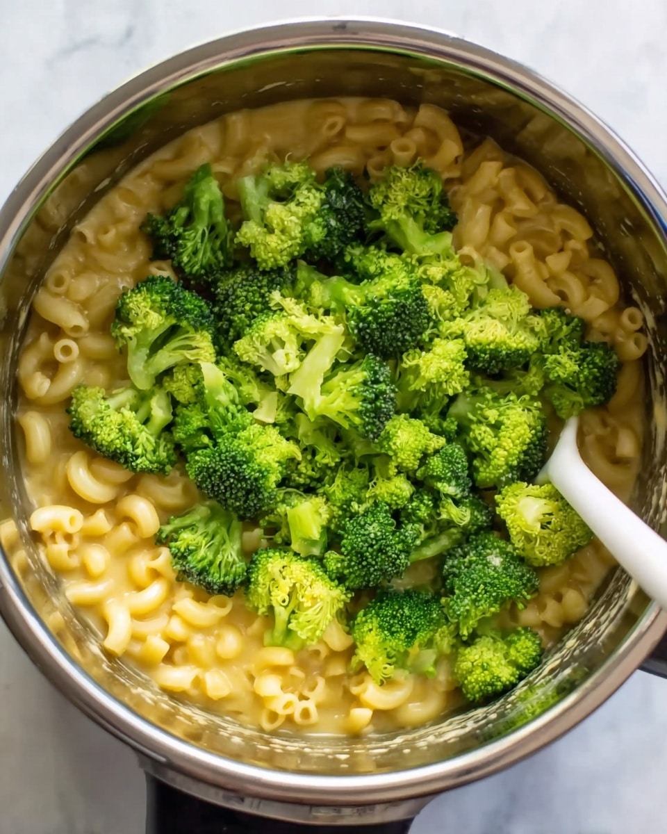 The image shows a close-up top view of a pot filled with cooked elbow macaroni pasta in a creamy yellow sauce. On top of the pasta, there is a generous layer of bright green broccoli florets that look fresh and slightly steamed. A white spoon is partially submerged in the pasta on the right side of the pot, with its handle resting against the broccoli. The pot sits on a white marbled surface that adds a clean background to the image. The colors mainly contrast between the yellow pasta, vibrant green broccoli, and shiny metallic pot. photo taken with an iphone --ar 4:5 --v 7