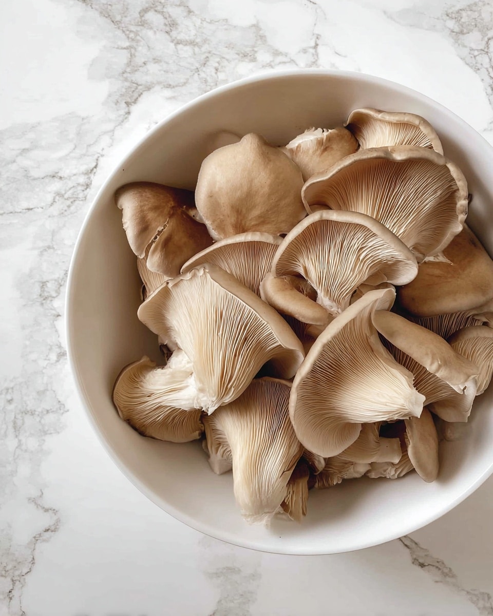 A white bowl filled with many oyster mushrooms that have a light brown color and smooth texture. The mushrooms are layered naturally, some showing the curved caps with faint cracks and the gills at the bottom. The bowl is placed on a white marbled surface with soft grey veins. photo taken with an iphone --ar 4:5 --v 7