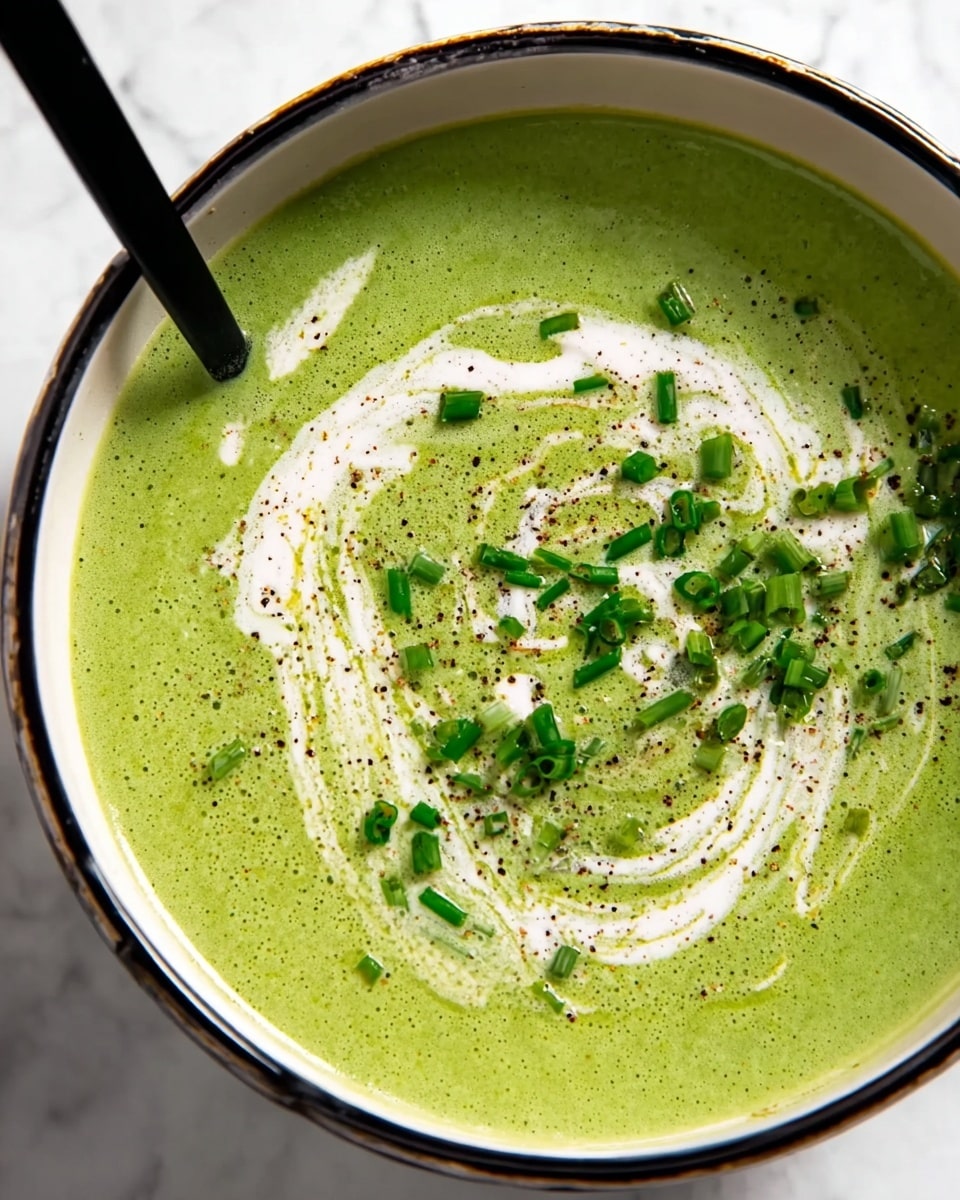 The image shows a close-up of a thick green soup with a smooth texture in a white bowl with a black rim. The soup has a swirl of white cream on top, creating a decorative pattern. Small green chopped chives are sprinkled in the center and a few are scattered around. There is a sprinkle of ground black pepper adding dark specks on the surface. A black spoon partially dips into the soup from the left side. The bowl sits on a white marbled texture photo taken with an iphone --ar 4:5 --v 7