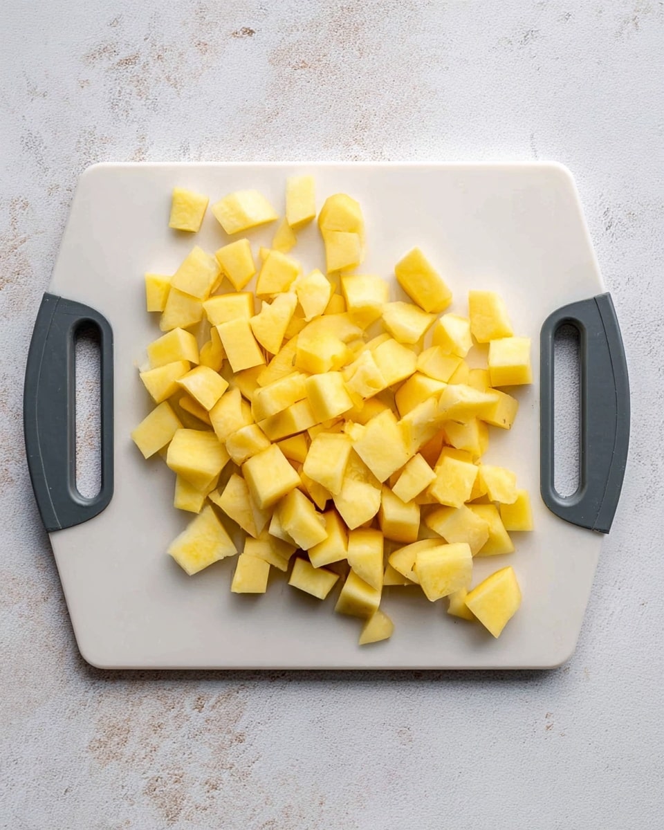 A white cutting board with dark gray handles holds many small yellow potato cubes scattered across its middle. The potatoes are cut into uneven small square and rectangular shapes with smooth textures. The cutting board is placed on a white marbled stone surface. The photo taken with an iphone --ar 4:5 --v 7