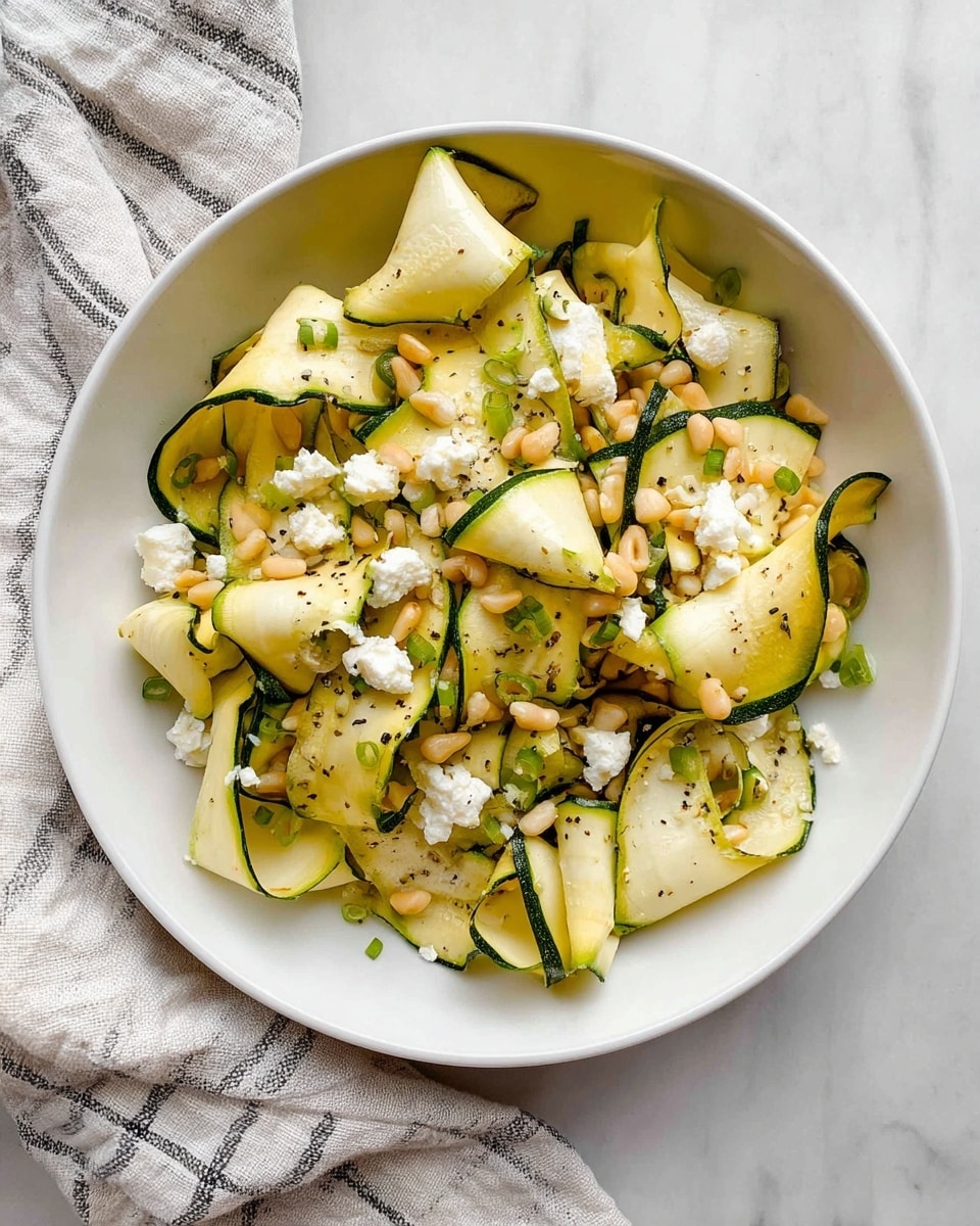 A white bowl filled with thin, folded ribbons of pale yellow zucchini with dark green edges, mixed with small light brown pine nuts and scattered chunks of white feta cheese. The zucchini ribbons have a soft, slightly shiny texture and are tossed with tiny green onion pieces and a few specks of black pepper on top. The bowl is placed on a white marbled surface with a soft striped cloth nearby. photo taken with an iphone --ar 4:5 --v 7