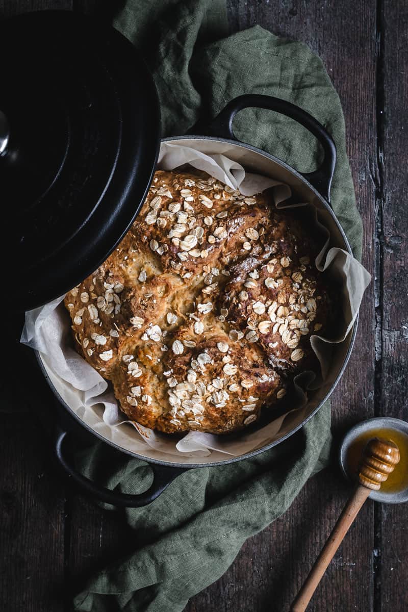 A round loaf of bread with a rough golden-brown crust sits in a black pot lined with white parchment paper. The bread top is sprinkled with light beige oat flakes, and the surface shows uneven, chunky cracks. The pot rests on a dark green cloth on a dark wooden table, with part of the black lid leaning on the left side of the pot. To the lower right, there is a small dish of honey with a wooden honey dipper nearby. The background is a white marbled texture. photo taken with an iphone --ar 4:5 --v 7