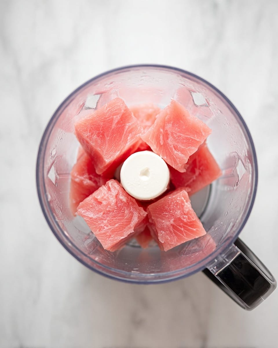 The image shows a transparent blender bowl filled with several thick, square pieces of fresh, pink fish placed evenly around a white blade in the center. The fish pieces have a smooth texture and are stacked in two layers, filling the bowl almost to the top. The blender bowl is placed on a white marbled surface, and the handle of the blender is visible on the right side. photo taken with an iphone --ar 4:5 --v 7