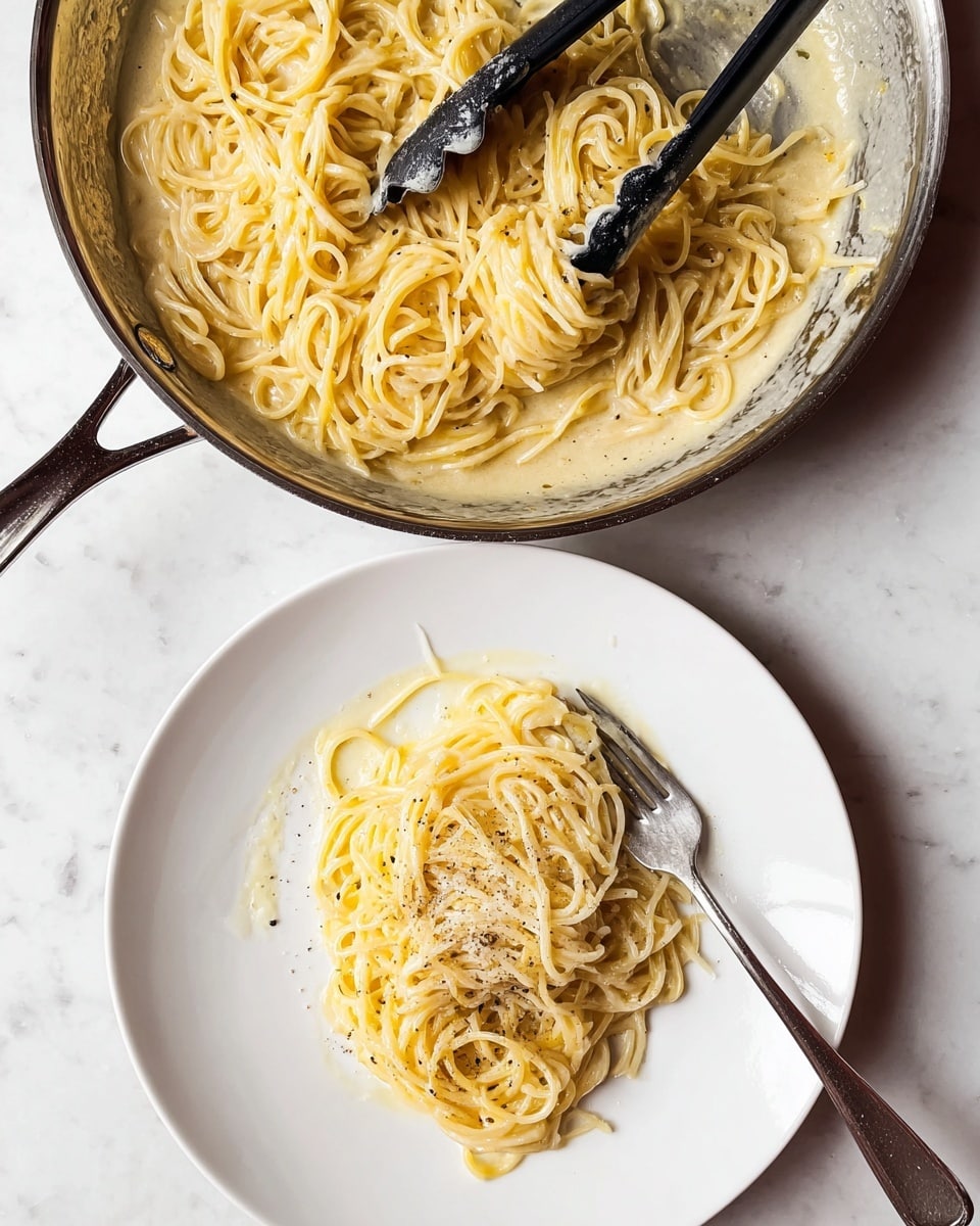 A white plate holds a small round serving of pasta with creamy light yellow sauce, sprinkled on one side with pale yellow grated cheese and black pepper. A fork twirls a portion of pasta on the right side of the plate. Above, a steel pan sits on a white marbled surface, holding more pasta mixed with creamy sauce and visible vanilla bean specks. Black tongs rest in the pan, partially submerged in the pasta. photo taken with an iphone --ar 4:5 --v 7