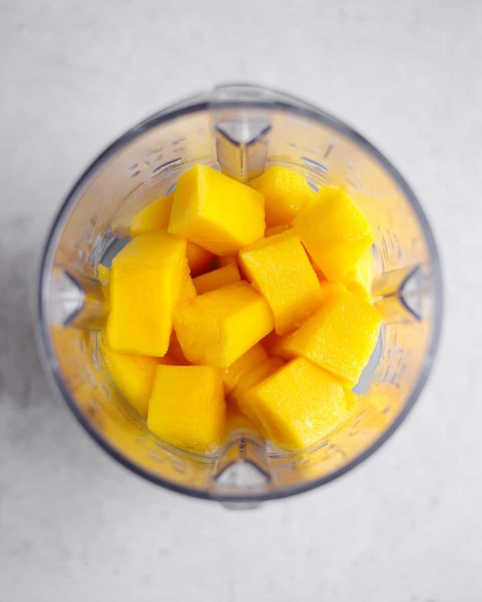 Inside a clear blender jar, there are bright yellow mango pieces that are cut into medium-sized cubes. The mango pieces fill almost half of the blender's height and are arranged unevenly. The blender jar has visible blades at the bottom. The background is a white marbled surface. photo taken with an iphone --ar 4:5 --v 7
