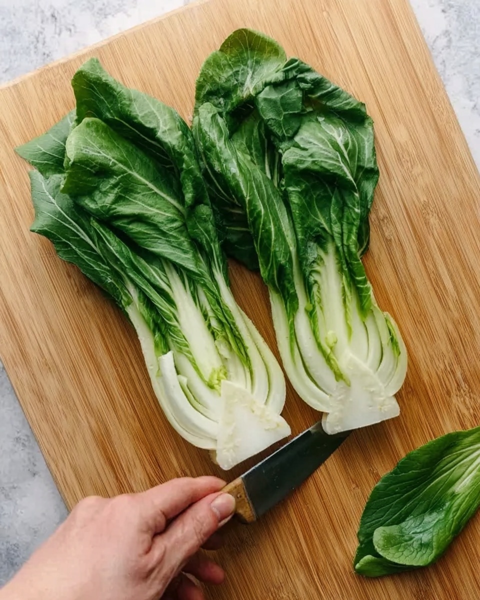 A woman's hand holding a fresh green and white bok choy cut in half, with the white stalks in the center and dark green leafy edges. The bok choy is placed on a light wooden cutting board, and a knife is positioned at the bottom right corner, slicing the leafy part. There is also a single green leaf lying on the board to the right. The background surface is replaced by a white marbled texture. photo taken with an iphone --ar 4:5 --v 7
