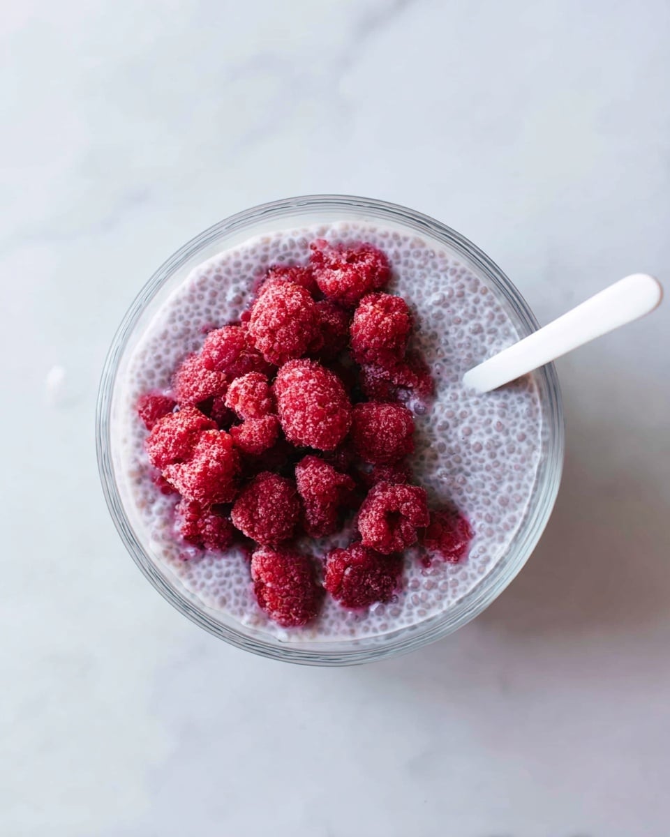 A clear glass bowl filled with a light purple, bubbly chia pudding base. On top, there is a layer of bright red frozen raspberries clustered in the center. A white spoon is placed inside the bowl on the right side, slightly touching the pudding. The bowl sits on a white marbled surface, creating a clean and fresh look. photo taken with an iphone --ar 4:5 --v 7