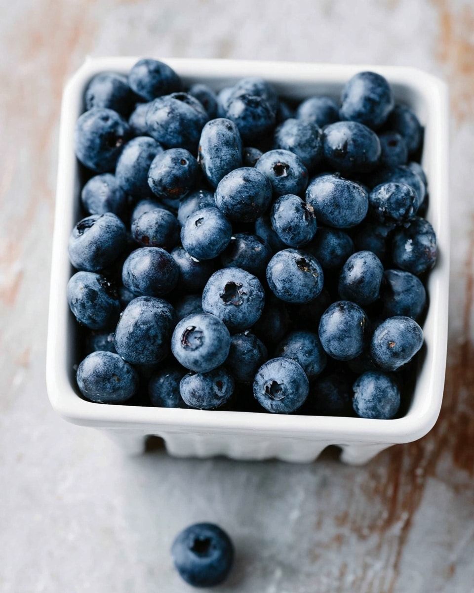 A white square container is filled with many fresh blueberries. The blueberries are deep blue with a natural matte finish, and they fill the container to the top with some berries resting on the edge. The background shows a white marbled texture that contrasts softly with the dark blueberries, highlighting their round shape and smooth texture. Photo taken with an iphone --ar 4:5 --v 7