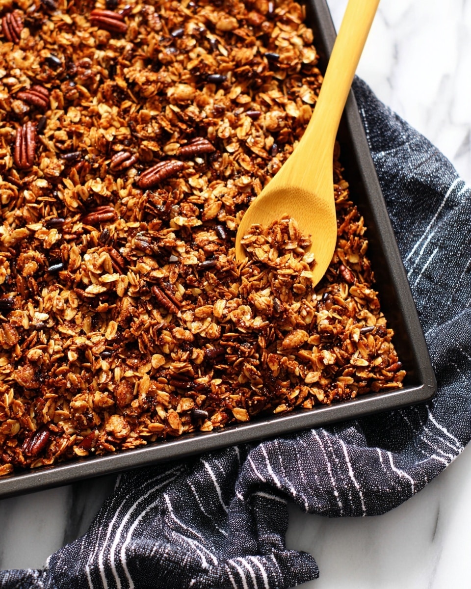 A close-up scene shows a dark baking tray filled with one thick, even layer of golden-brown toasted granola mixed with pecans and sunflower seeds, giving a rough and crunchy texture. A wooden spoon with a yellow handle rests on the tray’s right side, partly buried in the granola mix. The tray is placed on a white marbled surface with a black and white striped cloth partially underneath, adding contrast to the rich, warm colors of the granola photo taken with an iphone --ar 4:5 --v 7