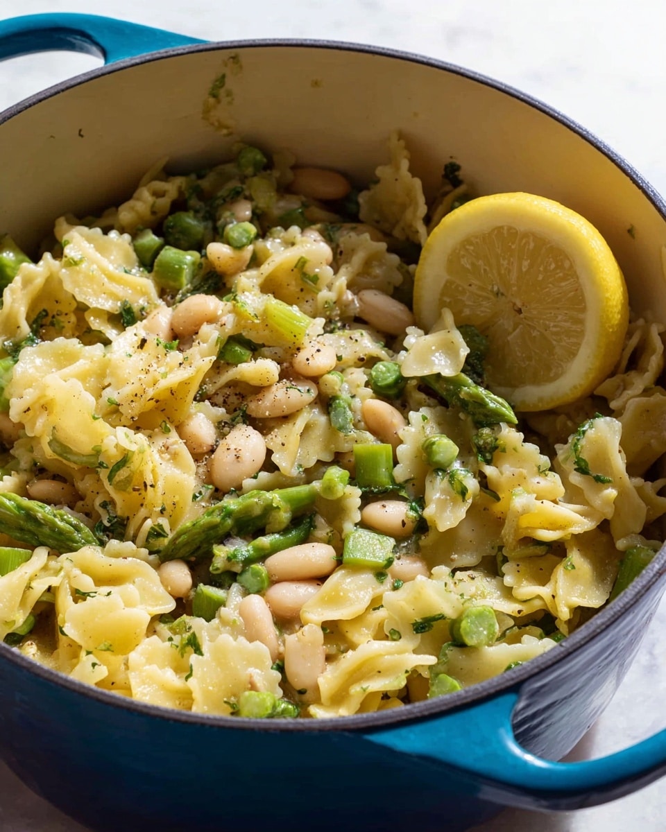 A close-up image of a blue pot filled with cooked pasta, chopped green asparagus pieces, white beans, and small bits of green herbs. The pasta has a short, ruffled shape and is light yellow. On the right side inside the pot, there is a slice of lemon resting on top of the mixture. The mixture looks moist with a slight gloss and is sprinkled lightly with black pepper. The pot sits on a white marbled surface in soft natural light. Photo taken with an iphone --ar 4:5 --v 7