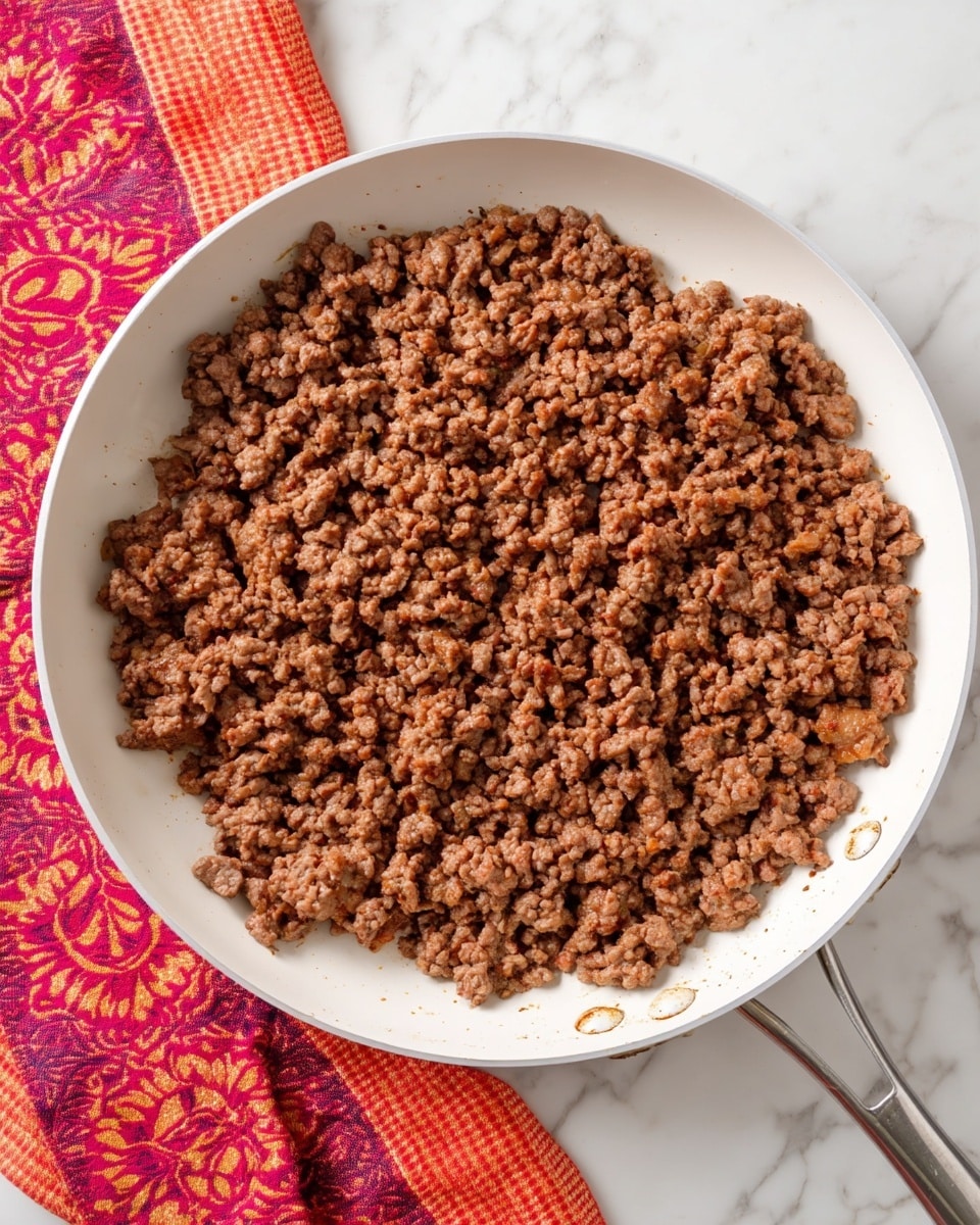A white frying pan filled with one layer of cooked ground meat that is brown and crumbly in texture, spread evenly across the pan. The pan is sitting on a white marbled surface with a red and orange patterned cloth partially visible on the left side. The pan handle extends out towards the bottom right corner of the image. Photo taken with an iphone --ar 4:5 --v 7