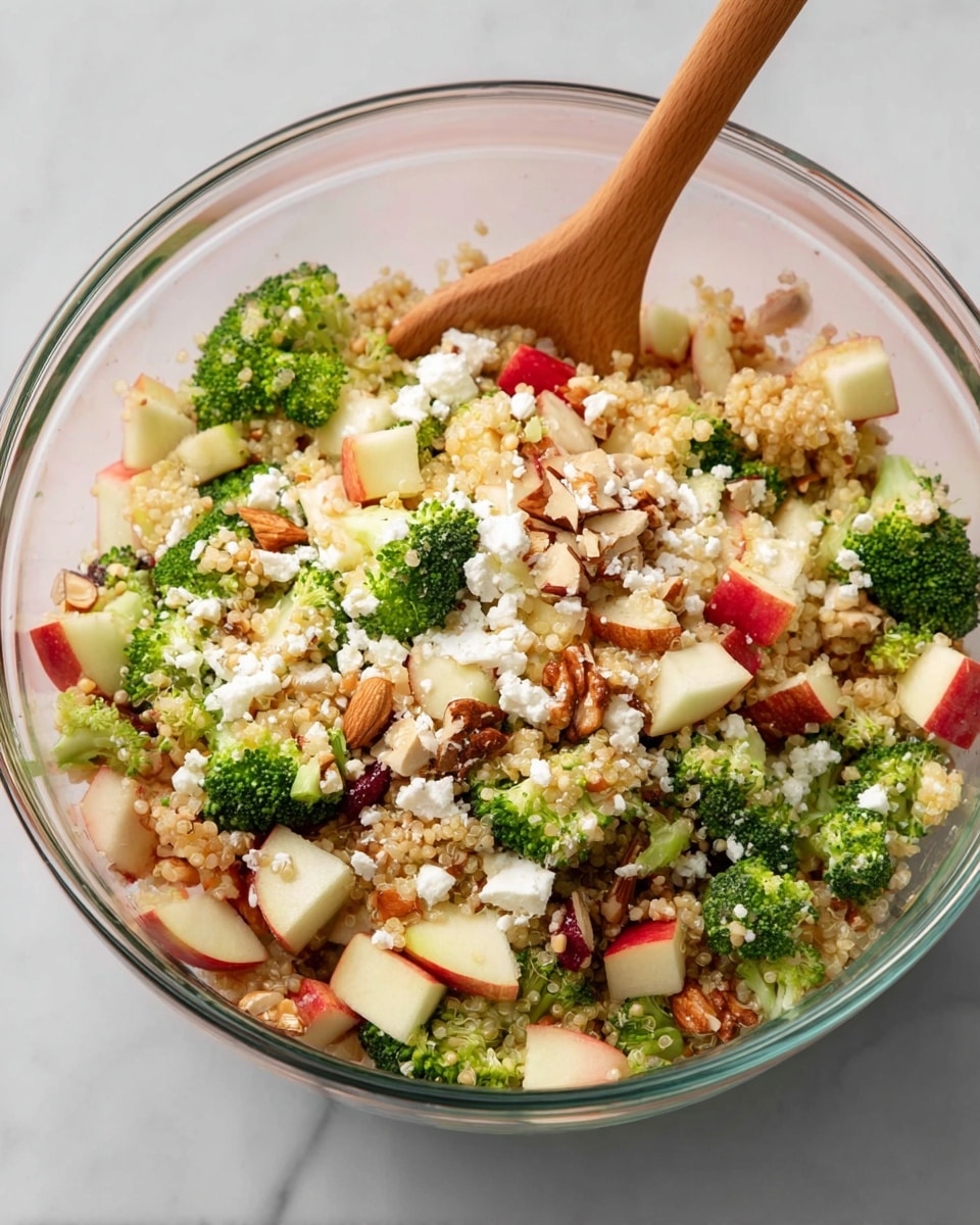 A clear glass bowl filled with a colorful salad sits on a white marbled surface. The salad has a base of light beige quinoa, mixed with green broccoli florets scattered evenly throughout. There are small cubes of red and white apple and pieces of light brown walnuts and almonds spread across the salad. White crumbled cheese is sprinkled on top, adding texture and contrast. A wooden spoon rests inside the bowl, partially buried in the salad. The photo taken with an iphone --ar 4:5 --v 7