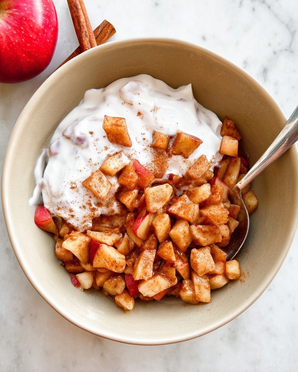 A beige, round bowl holds a mix of small, uneven apple pieces cooked to a soft texture with a warm brown color from cinnamon and spices. These apple chunks fill the bowl about halfway. On top, a thick layer of creamy white yogurt is spread unevenly, partly covering the apples. A silver spoon is placed inside the bowl on the right side, partly dipped into the mix. The bowl sits on a white marbled surface with a cinnamon stick nearby and a red apple partially visible in the top left corner. photo taken with an iphone --ar 4:5 --v 7