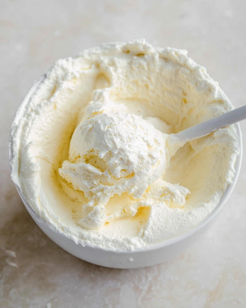 A white bowl filled with soft, fluffy white cream with a creamy, pale yellow scoop of ice cream or butter on top, showing smooth and airy textures. A white spoon is dipped into the creamy mixture from the top right side. The bowl is placed on a white marbled surface, with some cream spread near the edges inside the bowl. The image is close up and focuses on the creamy texture and colors photo taken with an iphone --ar 4:5 --v 7