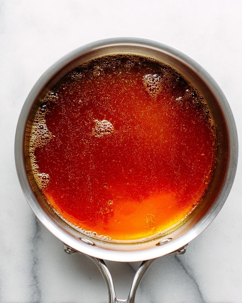 A silver metal pan filled with a single layer of amber to dark brown caramel liquid, showing a glossy surface with small bubbles and uneven texture. The pan rests on a white marbled surface, and the handles are visible in the lower right part of the image. The caramel color deepens more towards the bottom right side, giving a warm gradient effect. Photo taken with an iphone --ar 4:5 --v 7