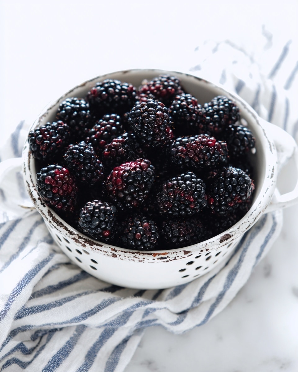 A white colander filled with dark blackberries that have a shiny, wet texture, showing deep black and dark red hues. The colander's edges have a worn, chipped white paint look revealing a rusty metal underneath. The colander sits on a folded white and blue striped cloth on a white marbled surface. Photo taken with an iphone --ar 4:5 --v 7