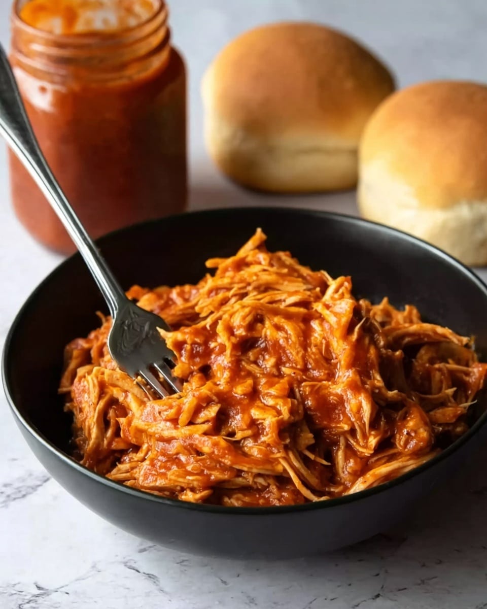 A black bowl filled with shredded chicken mixed with reddish-orange barbecue sauce, giving it a saucy and moist texture, with a fork stuck into the chicken on the left side of the bowl. Behind the bowl, there are two soft, rounded bread rolls and a glass jar filled with the same barbecue sauce, all placed on a white marbled surface. The focus is on the saucy shredded chicken, showing strands of meat coated well in the sauce. photo taken with an iphone --ar 4:5 --v 7