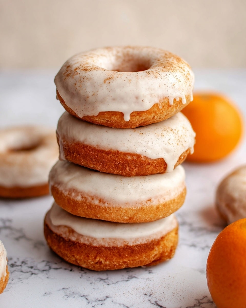 A stack of three round donuts with a light brown, fluffy texture is shown, each topped with a smooth, white glaze that slightly drips down the sides, with a light dusting of cinnamon or spice on top of the glaze. Around the stack, more similar donuts can be seen lying flat, and to the lower right, a small orange fruit is partially visible, all set against a white marbled surface. photo taken with an iphone --ar 4:5 --v 7