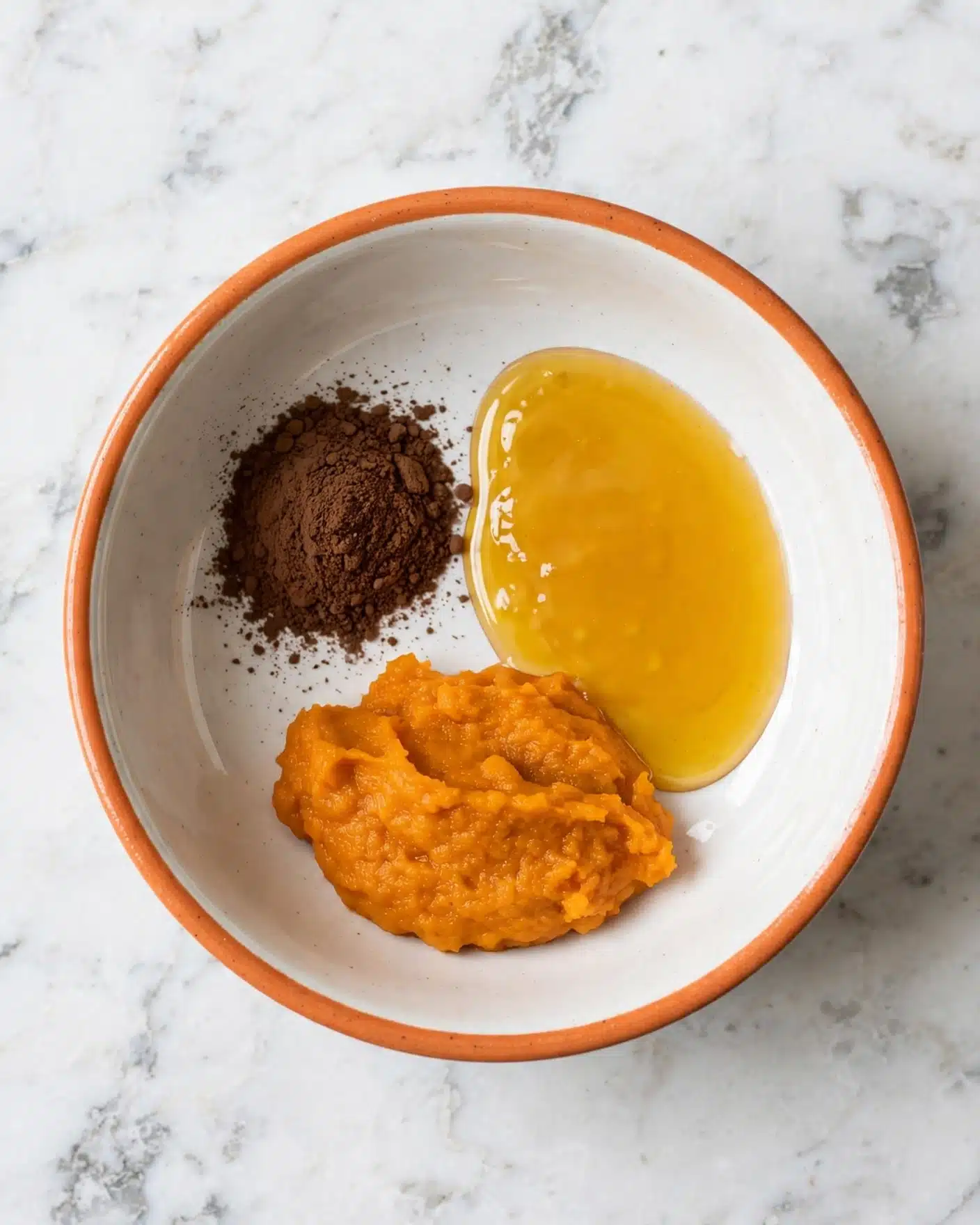Pumpkin Spice Milk Recipe 4 The image shows a white bowl with an orange rim on a white marbled surface, containing three separate ingredients inside: a small pile of dark brown powder on the left, a pool of golden liquid to the right, and a thick orange paste placed below them. The bowl is centered and viewed from above, highlighting the clear separation and different textures of each ingredient. Photo taken with an iphone --ar 4:5 --v 7