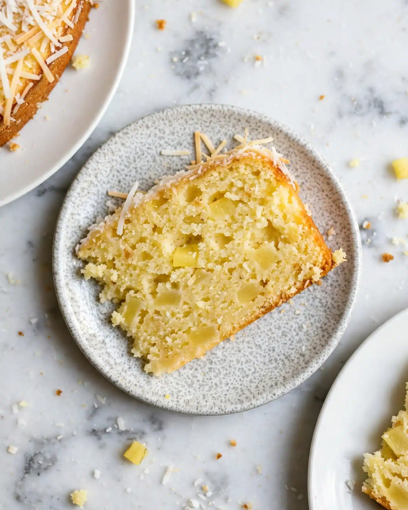 Vegan Mango Bread Recipe 5 The image shows a close-up of a single slice of yellow cake with a slightly crumbly texture, dotted with small light yellow fruit pieces inside. The top edge of the cake slice has a thin layer of shredded coconut. The slice is placed on a small round plate with a speckled, grayish-white textured pattern. Around the main plate, parts of two other white plates are visible, each holding slices of the same cake. The background is a white marbled surface, and some cake crumbs are scattered around. photo taken with an iphone --ar 4:5 --v 7