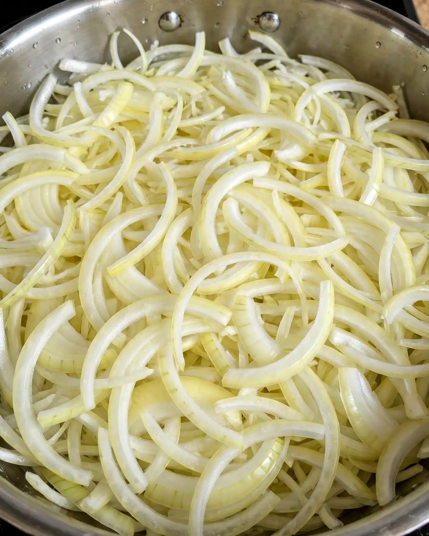 Creamy Caramelized Onion Pasta Recipe 4 A close-up view of many sliced raw onions inside a round metal pan. The onions are light yellow and white, thin and long with some curved pieces, filling the pan evenly with their soft, smooth texture. The pan has a shiny surface with a silver color and two small rivets visible at the top edge. The background is not visible, focusing on the fresh onions and the metal pan. photo taken with an iphone --ar 4:5 --v 7