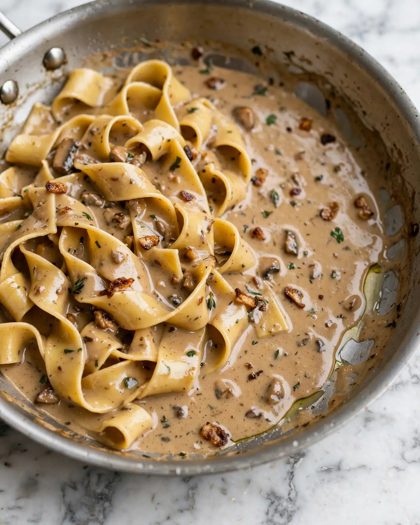Creamy Caramelized Onion Pasta Recipe 5 A close-up of a silver pan containing creamy pasta with a light brown sauce that coats thick, flat noodles evenly. The sauce has a smooth texture with small bits of herbs and darker browned ingredients mixed in, giving it subtle spots of darker brown. The pasta strands are twisted and layered loosely, resting mostly on the left side of the pan with some sauce pooling around the edges. The pan is placed on a white marbled surface. photo taken with an iphone --ar 4:5 --v 7