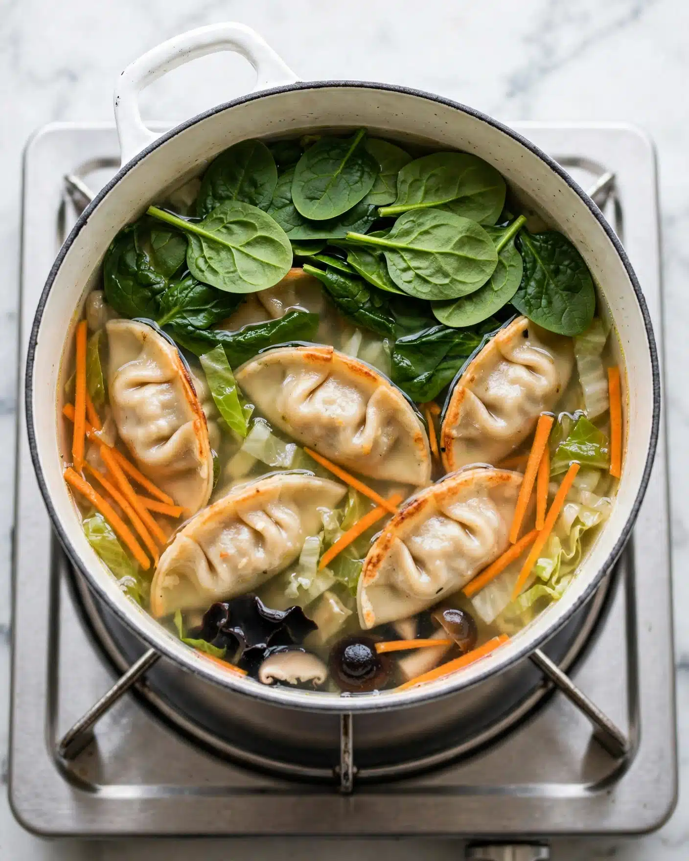 Potsticker Soup Recipe 4 A white pot filled with a clear broth soup, showing several half-moon shaped dumplings in light beige color lightly browned on the edges, sitting underneath a layer of bright green spinach leaves on top. Thin strips of orange carrot and bits of light green cabbage float in the soup around the dumplings, with some small black mushrooms visible near the bottom. The pot rests on a silver stove, with the background being white marbled texture. photo taken with an iphone --ar 4:5 --v 7
