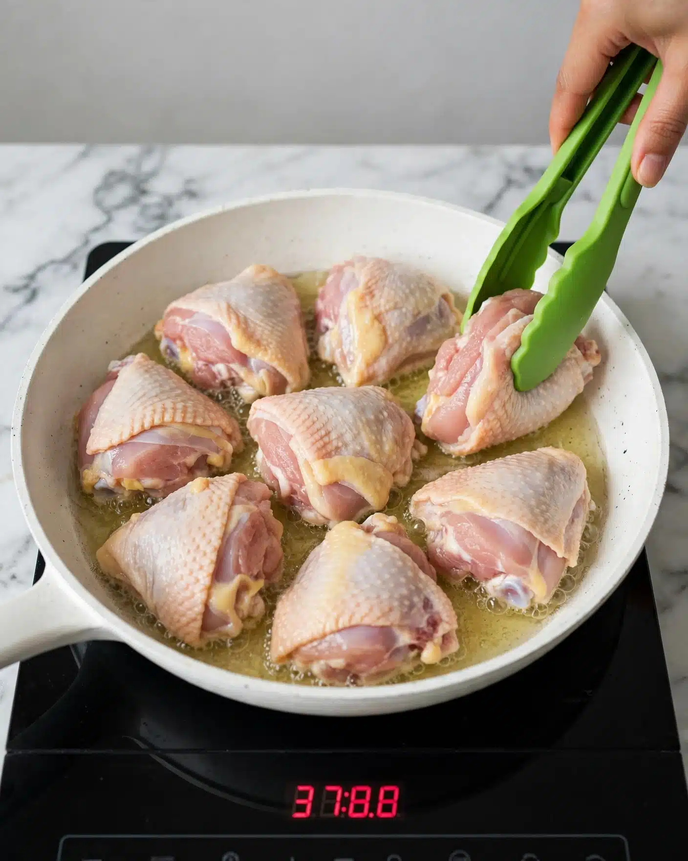 Easy One-Pan Lemon Parmesan Chicken & Orzo Recipe 4 A white pan with seven pieces of raw chicken skin side down cooking in oil, showing pale pink and yellow fat textures on each piece. A woman's hand is holding bright green tongs, placing one piece near the edge. The pan rests on a black induction cooktop with red digital numbers 37:8.8. The background is a white marbled surface and a light gray wall behind. Photo taken with an iphone --ar 4:5 --v 7