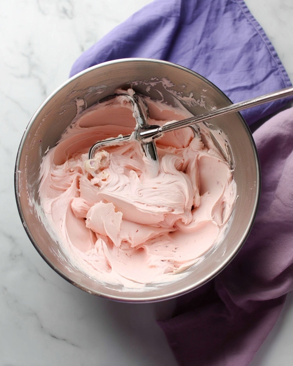 A shiny silver mixing bowl contains smooth, creamy pale pink frosting with soft swirls and folds showing a light, fluffy texture. Inside the bowl, a metal mixer paddle is resting among the frosting, which clings slightly to the sides of the bowl. The bowl is placed on a white marbled surface with a violet cloth partly underneath it. The overall feel is clean and simple with soft pastel color from the frosting. photo taken with an iphone --ar 4:5 --v 7