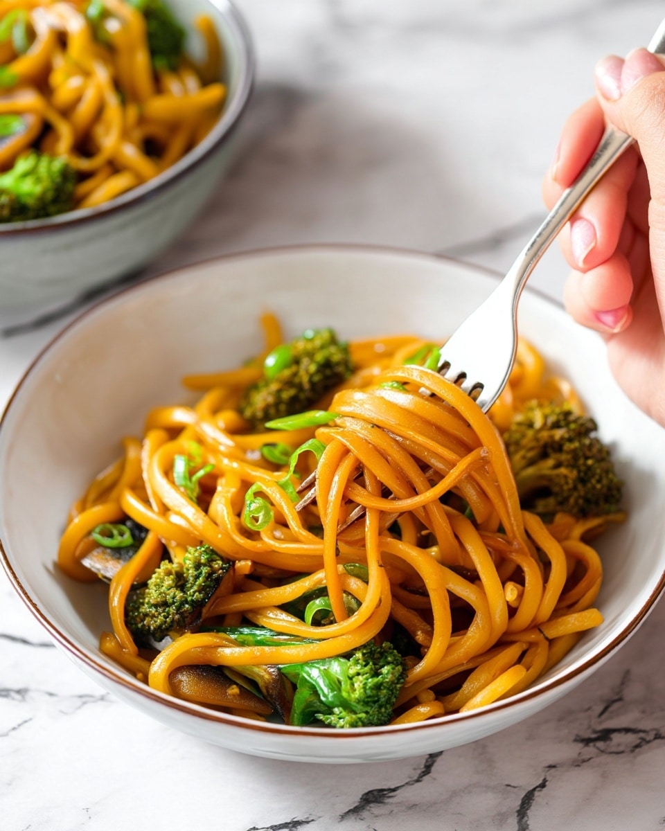A close-up of a bowl filled with thick, orange-colored noodles mixed with green broccoli pieces and small green chopped vegetables. The noodles have a smooth, shiny texture and are wrapped around a fork held by a woman's hand on the right side. The white bowl with a thin dark rim sits on a white marbled surface. In the blurry background, there is another bowl with a similar noodle dish. photo taken with an iphone --ar 4:5 --v 7