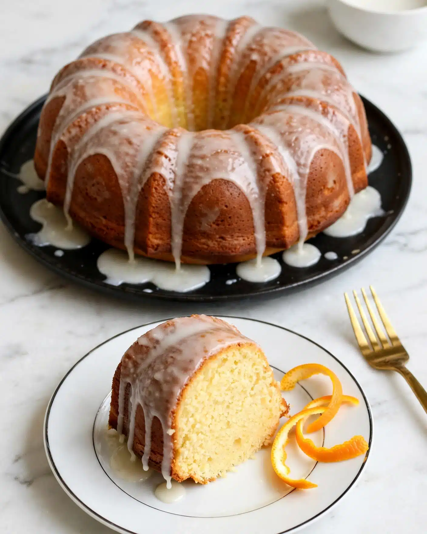 A thick slice of yellow bundt cake with a smooth, light brown crust is placed on a white plate with a thin black rim; the cake slice is topped with a pale, glossy glaze that drips slightly down its sides. Thin orange peel twists rest beside the slice on the plate. In the background, a bundt cake with the same yellow inside, brown crust, and light glaze sits on a black plate with some glaze pooled underneath it. The whole scene is set on a white marbled surface with a gold fork visible to the side. Photo taken with an iphone --ar 4:5 --v 7