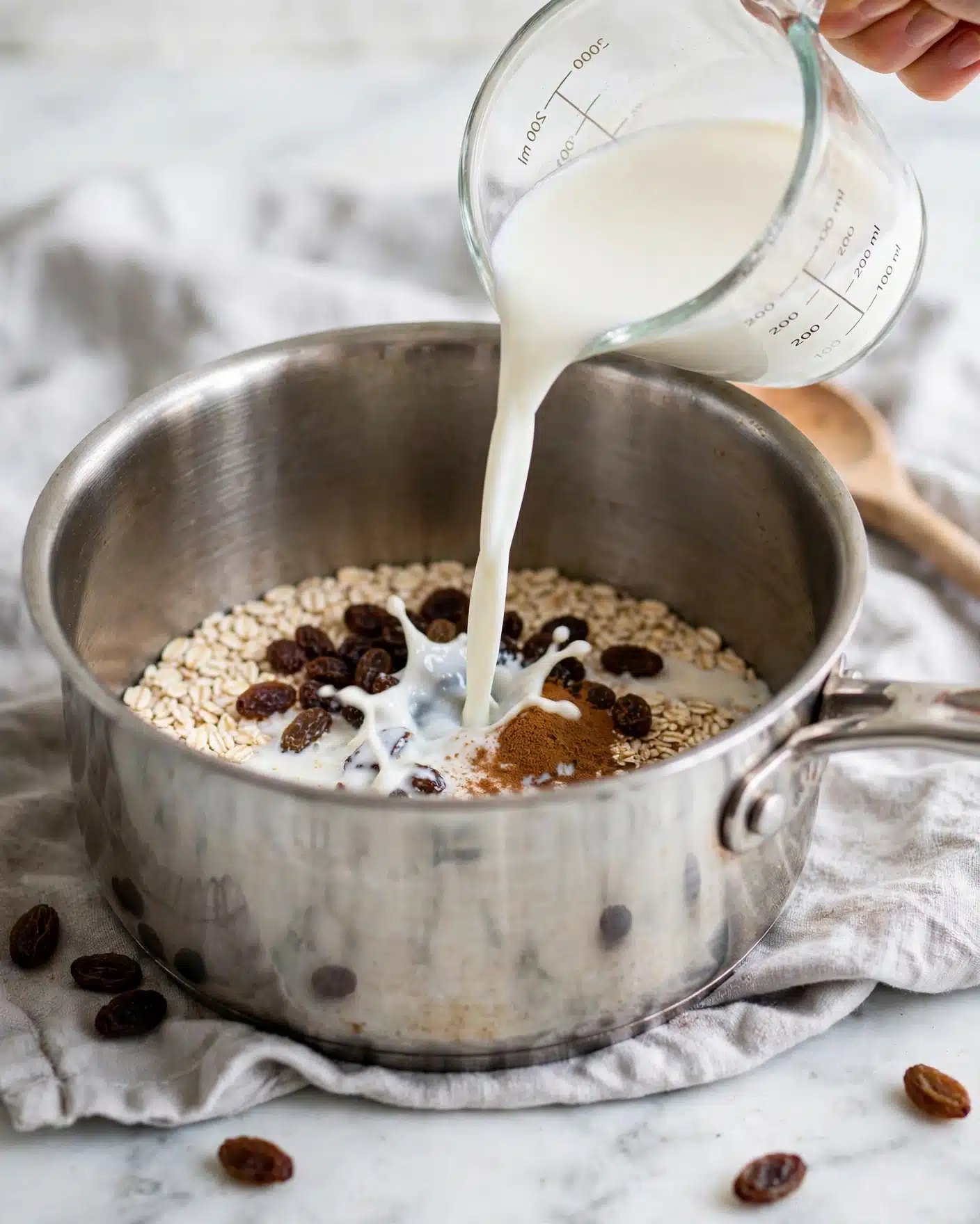 Instant Pot Rice Pudding Recipe 4 A close-up view of a shiny steel pot with a mixture inside consisting of white grains on the bottom, topped with some dark brown raisins, and a small pile of brown powder, all partially covered by a white liquid being poured from a clear measuring cup into the pot. The pot sits on a soft gray cloth, which lies on a white marbled surface. In the slightly blurred background, a wooden spoon handle is visible and a woman's hand is gently holding the measuring cup. photo taken with an iphone --ar 4:5 --v 7