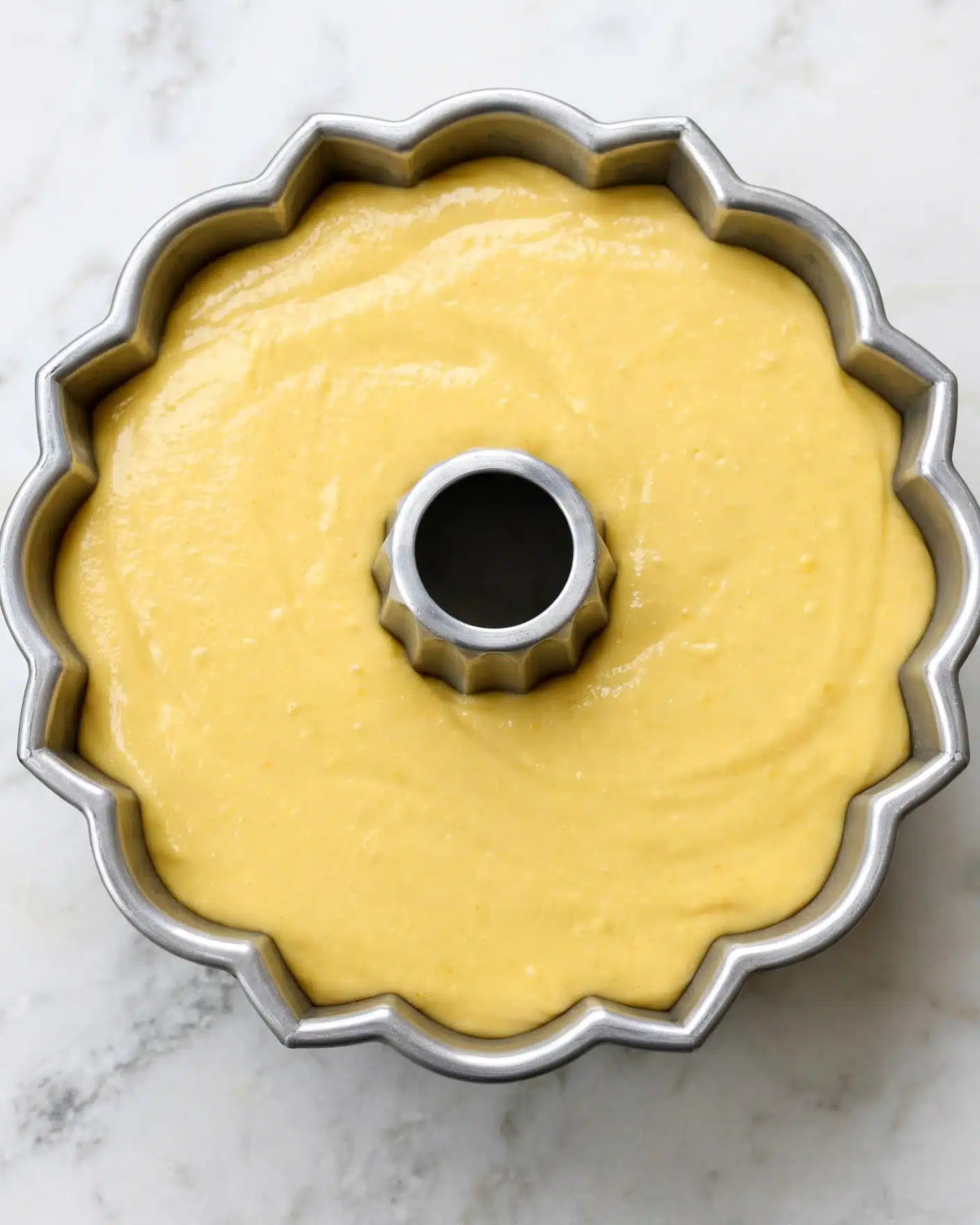 The image shows a metal Bundt pan filled with smooth, thick, yellow cake batter. The batter is spread evenly and smooth on top, filling the pan almost to the brim. The pan has fluted, tall sides and a central tube in the middle. The pan sits on a white marbled surface, and there are no other objects in the scene. Photo taken with an iphone --ar 4:5 --v 7