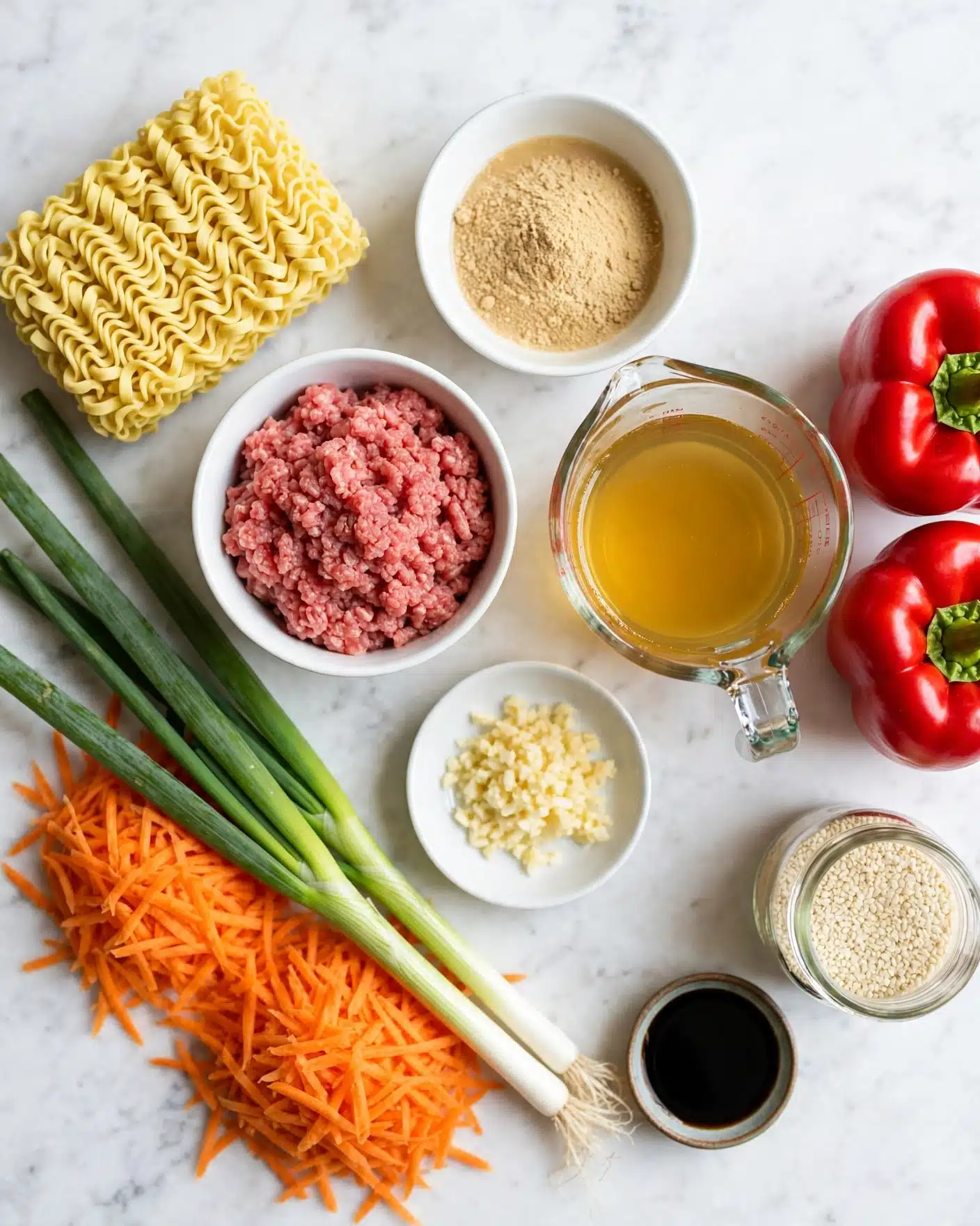 A flat lay image on a white marbled surface shows various ingredients arranged neatly. On the left side, there are uncooked yellow ramen noodles with a block shape and a wavy texture. Nearby, three green onions with long white roots and green tops stretch diagonally across the image, resting on piles of thin, bright orange shredded carrots. In the center-left, a white bowl holds a chunk of raw ground meat with a pinkish-red color and a rough texture. Right next to it is a small white bowl filled with light tan powder, resembling ground ginger or powdered broth. Below that bowl, another small white dish contains finely chopped garlic in a pale yellow shade. To the right of these bowls, a clear glass measuring cup holds a golden-yellow liquid broth. On the far right, two plump, shiny red bell peppers sit side by side. A small glass jar with white sesame seeds and a tiny round dish of a black liquid, probably soy sauce, complete the setup. Photo taken with an iphone --ar 4:5 --v 7