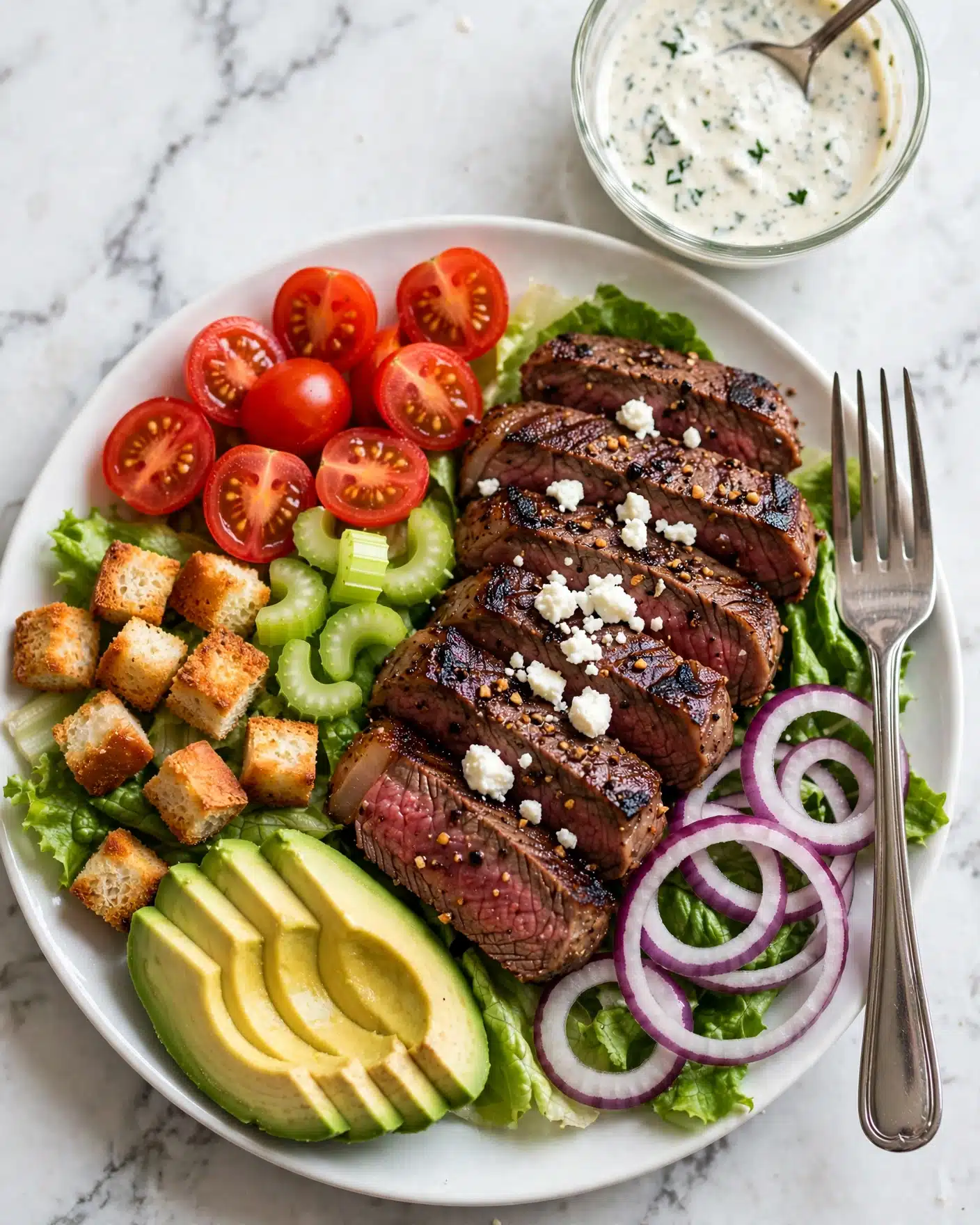 Black n' Blue Grilled Steak Salad Recipe 5 A white plate on a white marbled surface holds a colorful salad arranged in separate sections: bright red halved cherry tomatoes on the top left, golden-brown croutons below them on a bed of green lettuce, yellow-green avocado slices fanned out on the bottom left, crisp light green celery pieces near the center, and thin slices of grilled dark brown steak layered in the middle with small white cheese crumbles on top. To the right of the steak, there are rings of purple-red onion sitting on more lettuce, and a silver fork rests at the right edge of the plate. Above the plate, a small clear glass bowl of creamy white dressing with some green herbs and a spoon inside is visible. Photo taken with an iphone --ar 4:5 --v 7
