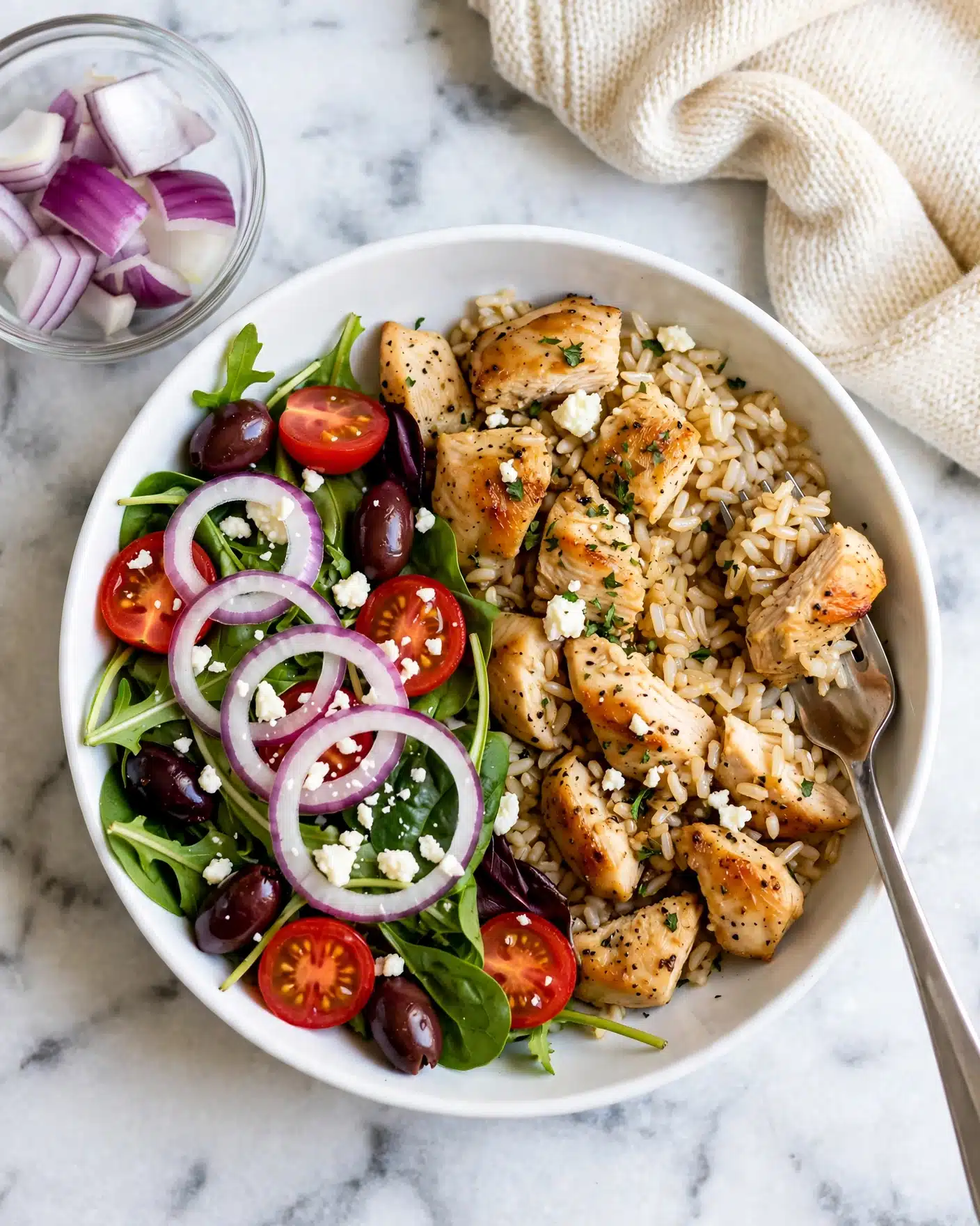 Instant Pot Greek Chicken and Rice Recipe 5 A white bowl is filled with two main parts: on the right side there is light brown rice mixed with small pieces of grilled light golden chicken, sprinkled with small white cheese crumbles and bits of green herbs; on the left side there is a fresh green salad made of mixed leafy greens, sliced red cherry tomatoes, dark purple olives, and thin pale purple onion rings, also topped with white crumbles of cheese. A silver fork holds a bite of rice and chicken on the right side of the bowl. To the upper left of the bowl, there is a small clear glass bowl with chunks of purple and white onion. The bowl and onion bowl are placed on a white marbled surface with a cream-colored knitted cloth at the top right corner. photo taken with an iphone --ar 4:5 --v 7