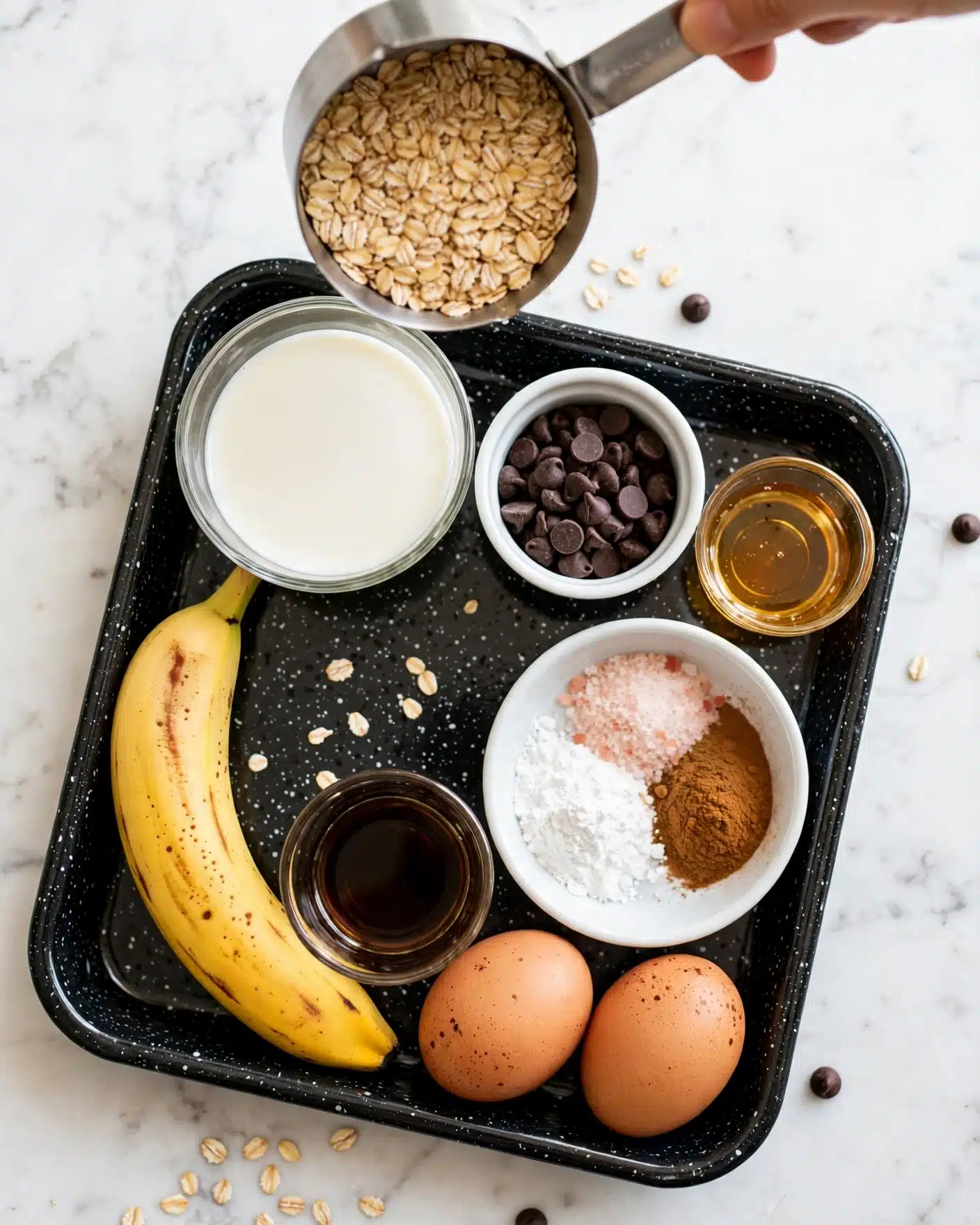 The image shows a small black speckled tray on a white marbled surface. Inside the tray, from top left going clockwise, there is a clear glass bowl filled with white milk, a white bowl holding dark brown chocolate chips, a small clear glass with honey, a small white bowl with three different powders (white baking powder, light pink salt, and brown cinnamon) arranged in separate piles, two brown eggs at the bottom right corner, a clear glass with dark liquid (likely vanilla extract), a ripe yellow banana with brown spots on the left side, and a metal measuring cup filled with rolled oats held above the tray. The colors range from white and light brown to dark brown and yellow, with simple and natural textures. Photo taken with an iphone --ar 4:5 --v 7