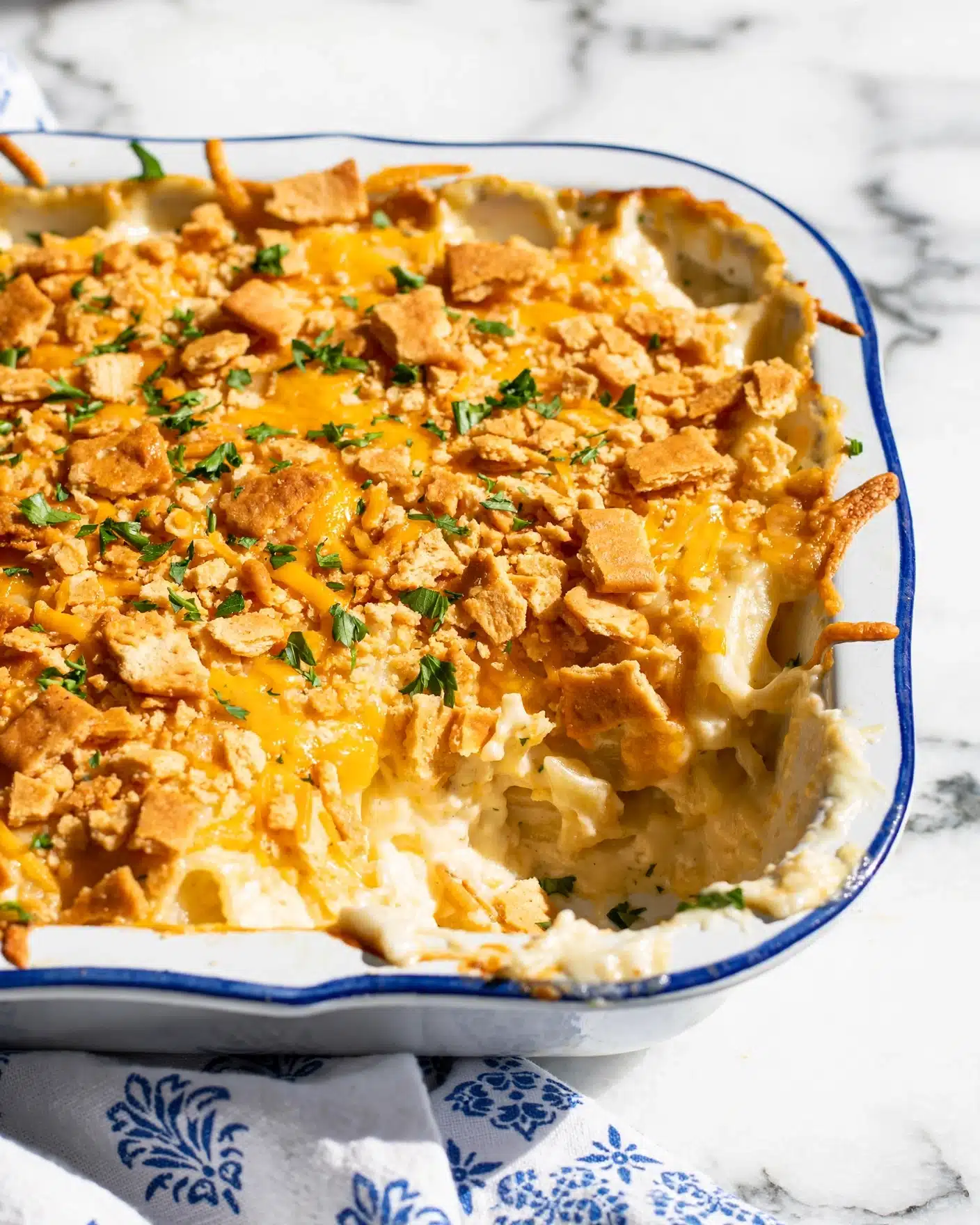 A close-up of a casserole dish showing a rich layered baked dish with a golden brown and slightly crispy top layer made of crushed crackers and shredded cheddar cheese, sprinkled with fresh green parsley bits. The dish is white with a blue edge, and the texture looks creamy under the crispy topping. The background is a white marbled texture with a white cloth with blue patterns under the dish. photo taken with an iphone --ar 4:5 --v 7