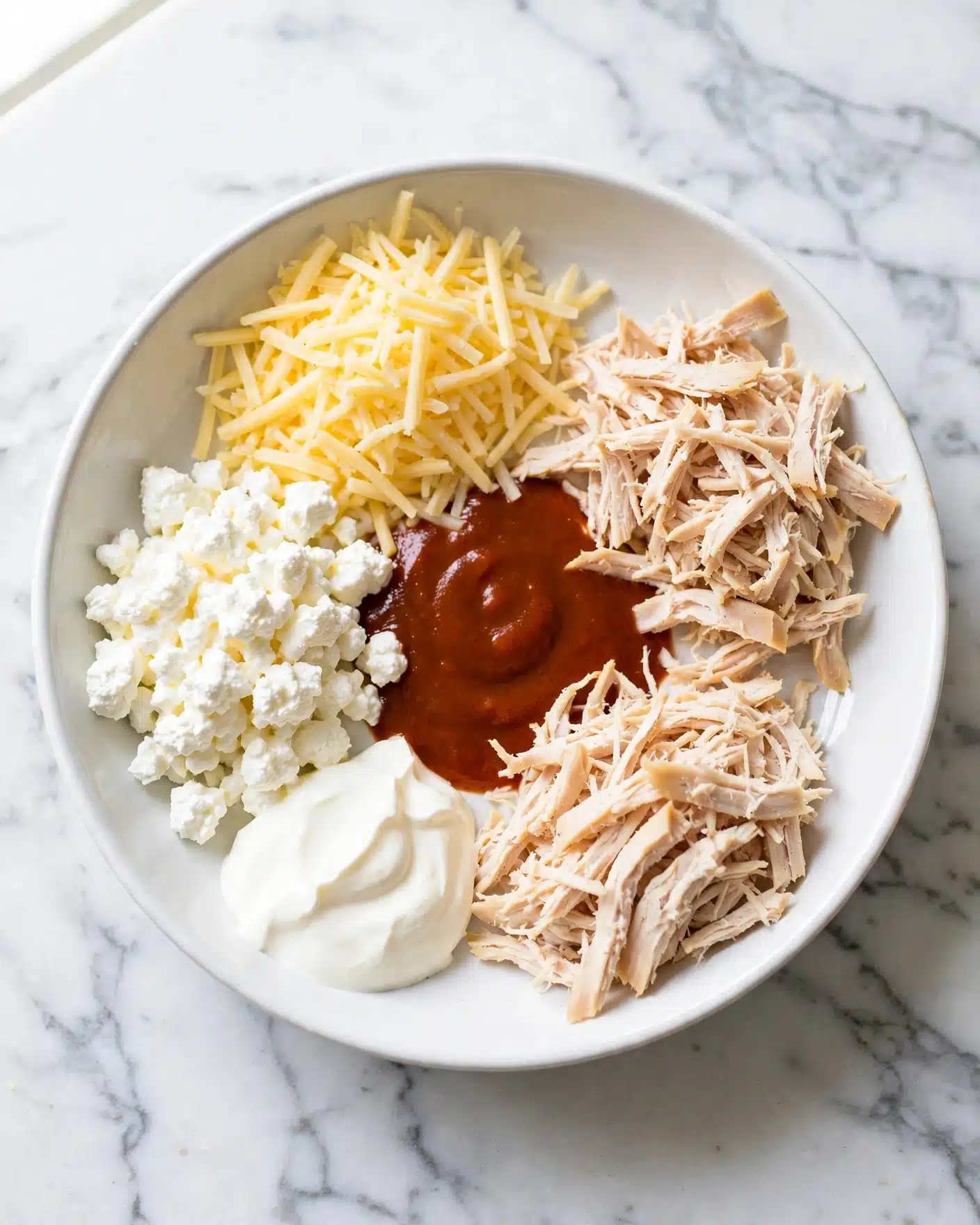 Healthy Buffalo Chicken Dip Recipe 4 A top view of a white bowl on a white marbled surface with five separate layers inside. The first layer is shredded pale yellow cheese piled on the top left. Below it, there is a cluster of white cottage cheese with a lumpy texture. On the bottom left, there is a dollop of smooth, creamy white sour cream. The center has a reddish-brown sauce spread in the middle. To the right of the sauce, there are two piles of shredded light beige chicken, one above and one below the sauce. Photo taken with an iphone --ar 4:5 --v 7