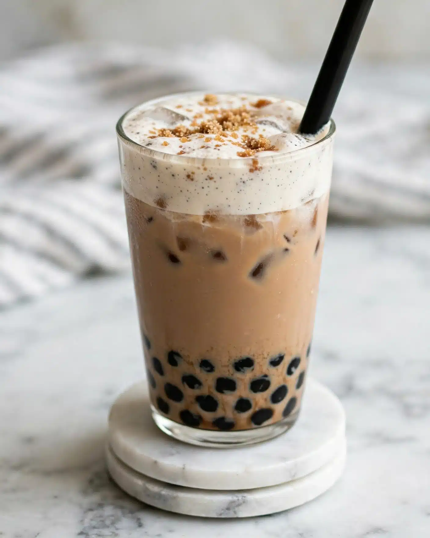 A clear glass filled with a light brown iced drink with visible black tapioca pearls settled at the bottom layer, topped with a thick, creamy white foam layer sprinkled with brown sugar bits on top, and a thick black straw inserted on one side; the glass sits on two stacked white marble coasters with a soft-focused white marbled surface in the background and a blurred white and gray striped cloth behind. photo taken with an iphone --ar 4:5 --v 7