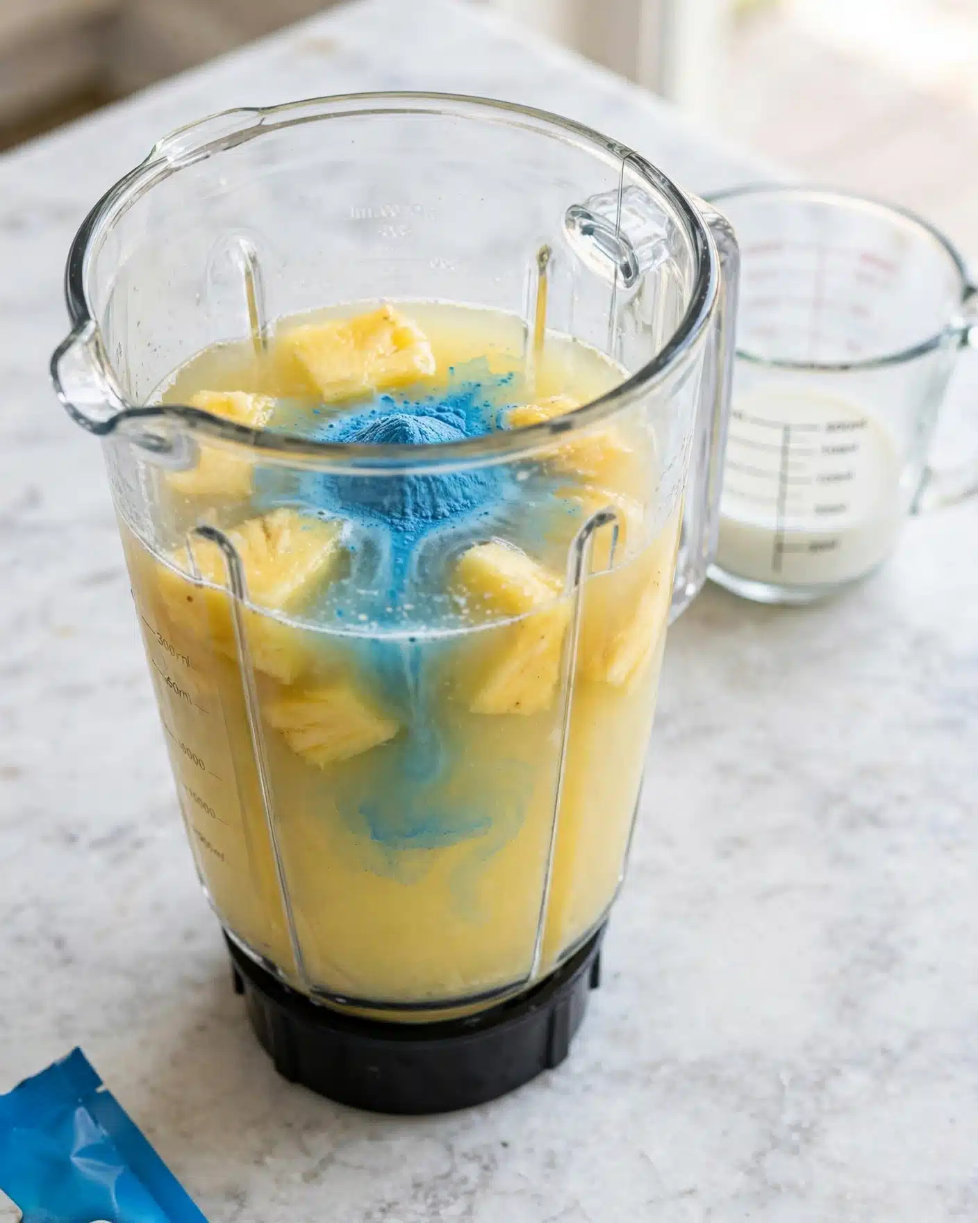 Blue Milk (Star Wars Inspired) Recipe 4 A clear blender container filled with a light yellow liquid mixture with chunks of yellow fruit, likely pineapple, floating inside. On top of the fruit and liquid, there is a small pile of bright blue powder that is dissolving and spreading into the mixture. The blender is placed on a white marbled surface, with a clear measuring cup containing a small amount of white liquid visible to the upper right. Part of a blue package is visible at the bottom left corner. Photo taken with an iphone --ar 4:5 --v 7