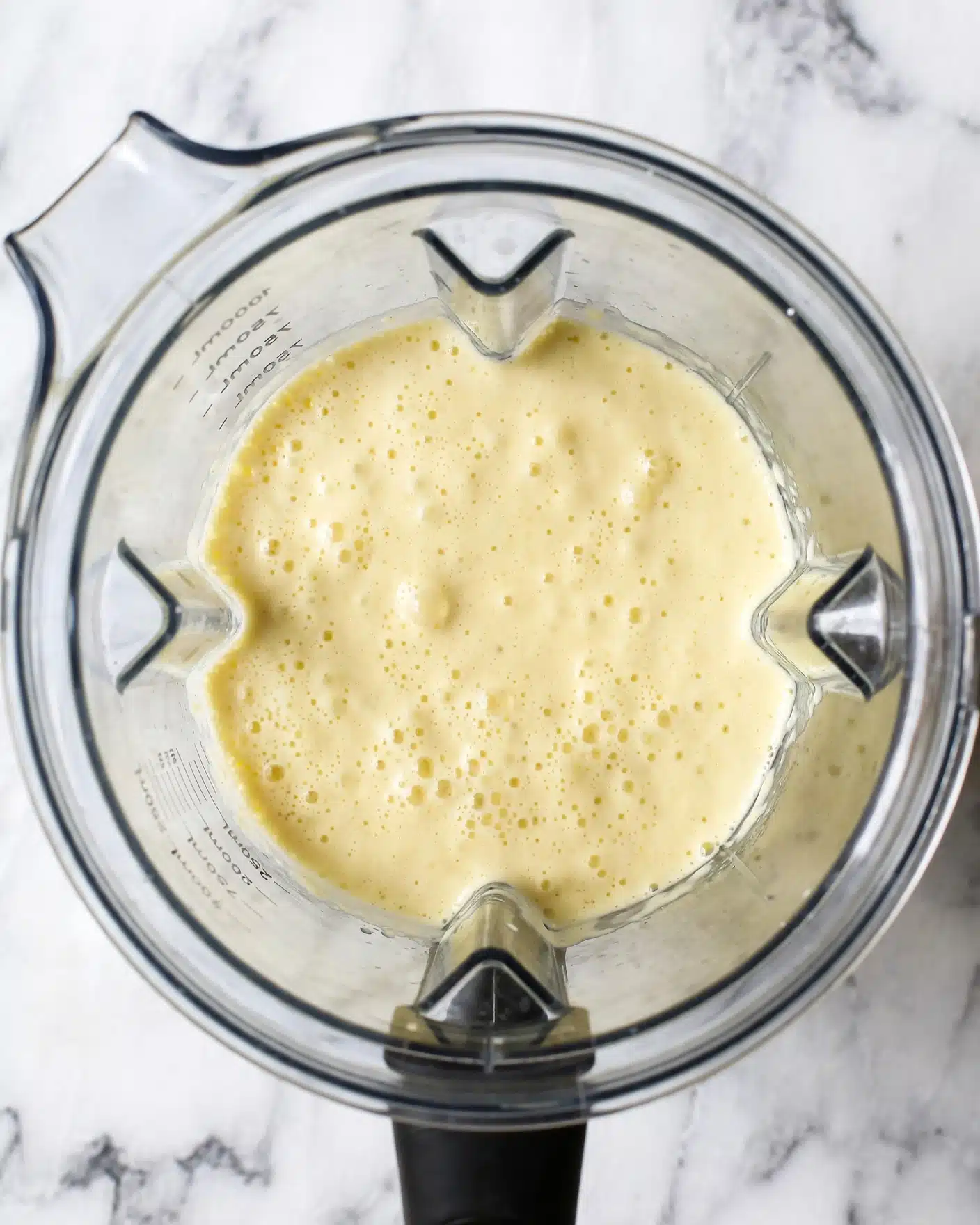 A clear blender container filled with a smooth, pale yellow creamy mixture, slightly bubbly with tiny air holes on the surface, sitting on a white marbled texture background, viewed from above. photo taken with an iphone --ar 4:5 --v 7