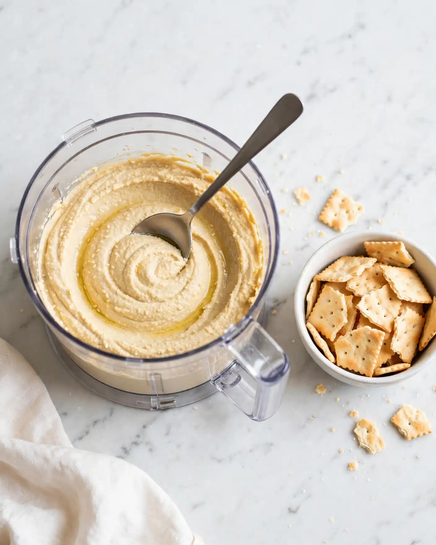 Spicy Piri Piri Hummus Recipe 4 A clear food processor bowl filled with a creamy, light beige hummus-like spread smooth in texture with gentle swirls made by a spoon resting inside. Next to it, a white bowl holds several broken pieces of light golden-brown crackers with a slightly rough surface. The scene is set on a white marbled texture background with a soft off-white cloth partially visible in the bottom left corner. Photo taken with an iphone --ar 4:5 --v 7
