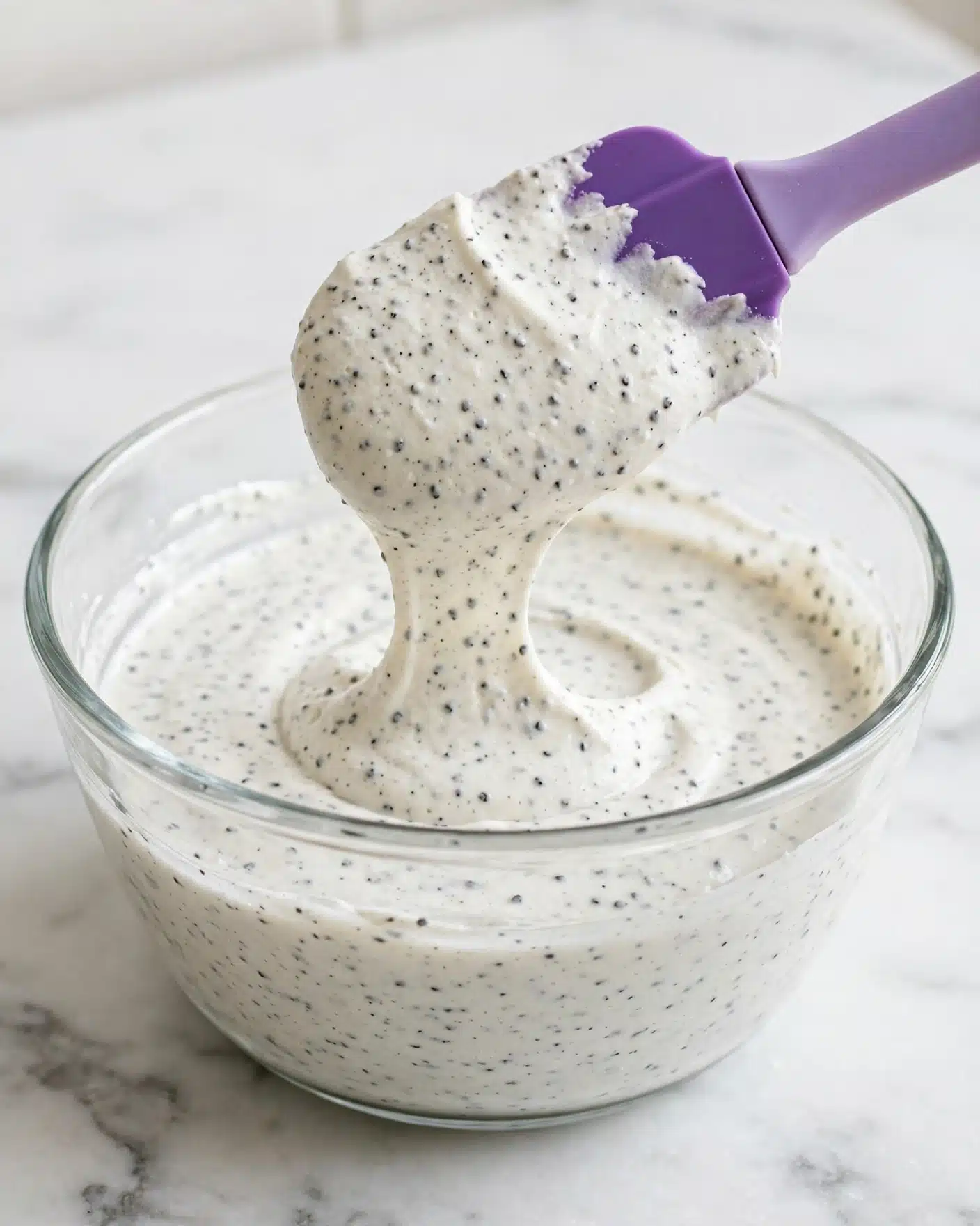 A clear glass bowl filled with a creamy white mixture that has a thick, smooth texture with small black specks evenly spread throughout. A purple silicone spatula lifts a scoop of the mixture above the bowl, showing its soft, flowing consistency. The background surface is white marbled, creating a clean and bright setting. Photo taken with an iphone --ar 4:5 --v 7
