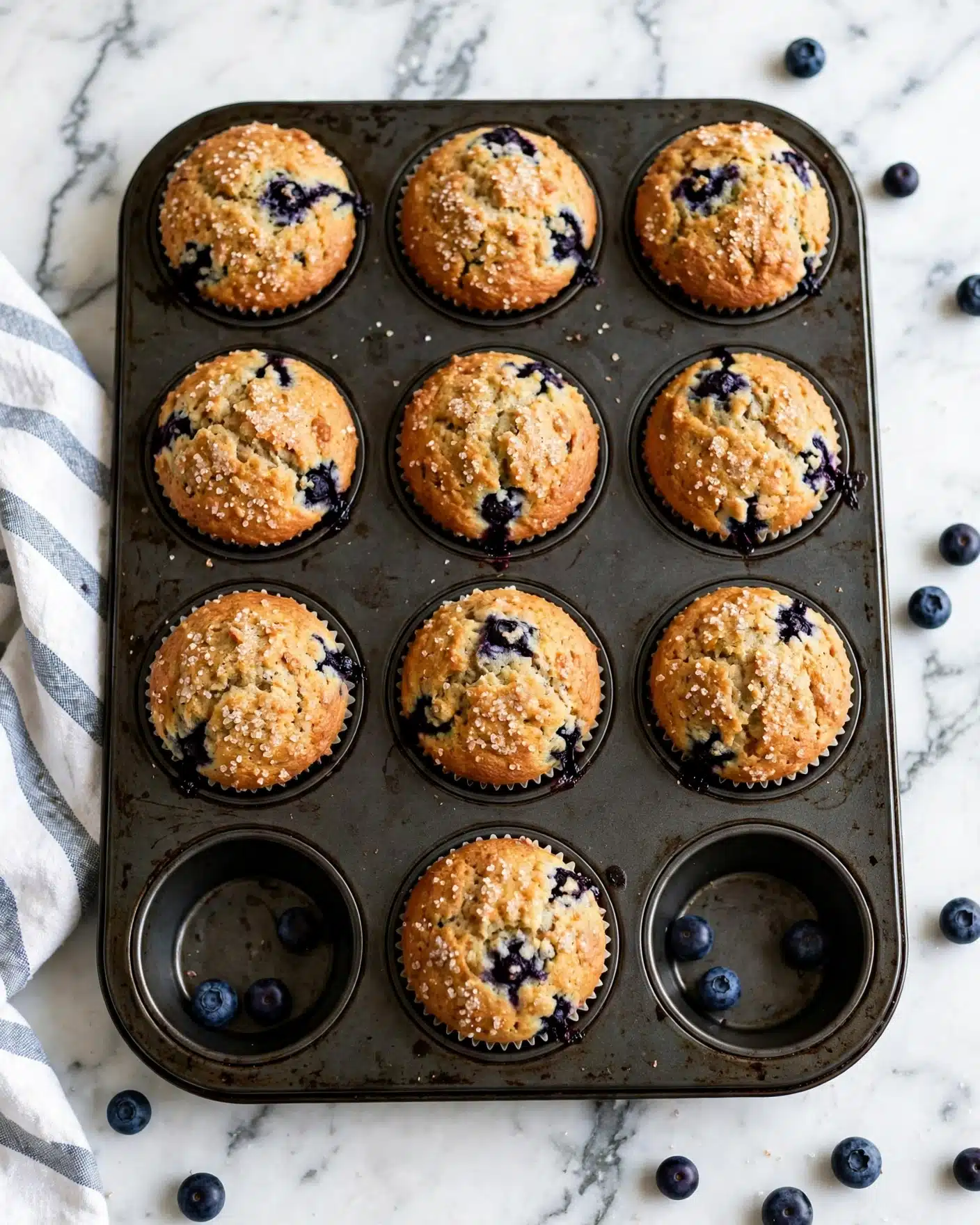 A dark metal muffin tray with eight golden brown blueberry muffins, each muffin showing several dark purple blueberry spots on top and some peeking from the sides inside white liners. The muffins have a slightly rough textured top with some sugar sparkle. Scattered fresh blueberries are placed in the empty muffin slots and around the tray, resting on a white marbled surface. A white and blue striped cloth is partially visible on the left side of the image. photo taken with an iphone --ar 4:5 --v 7