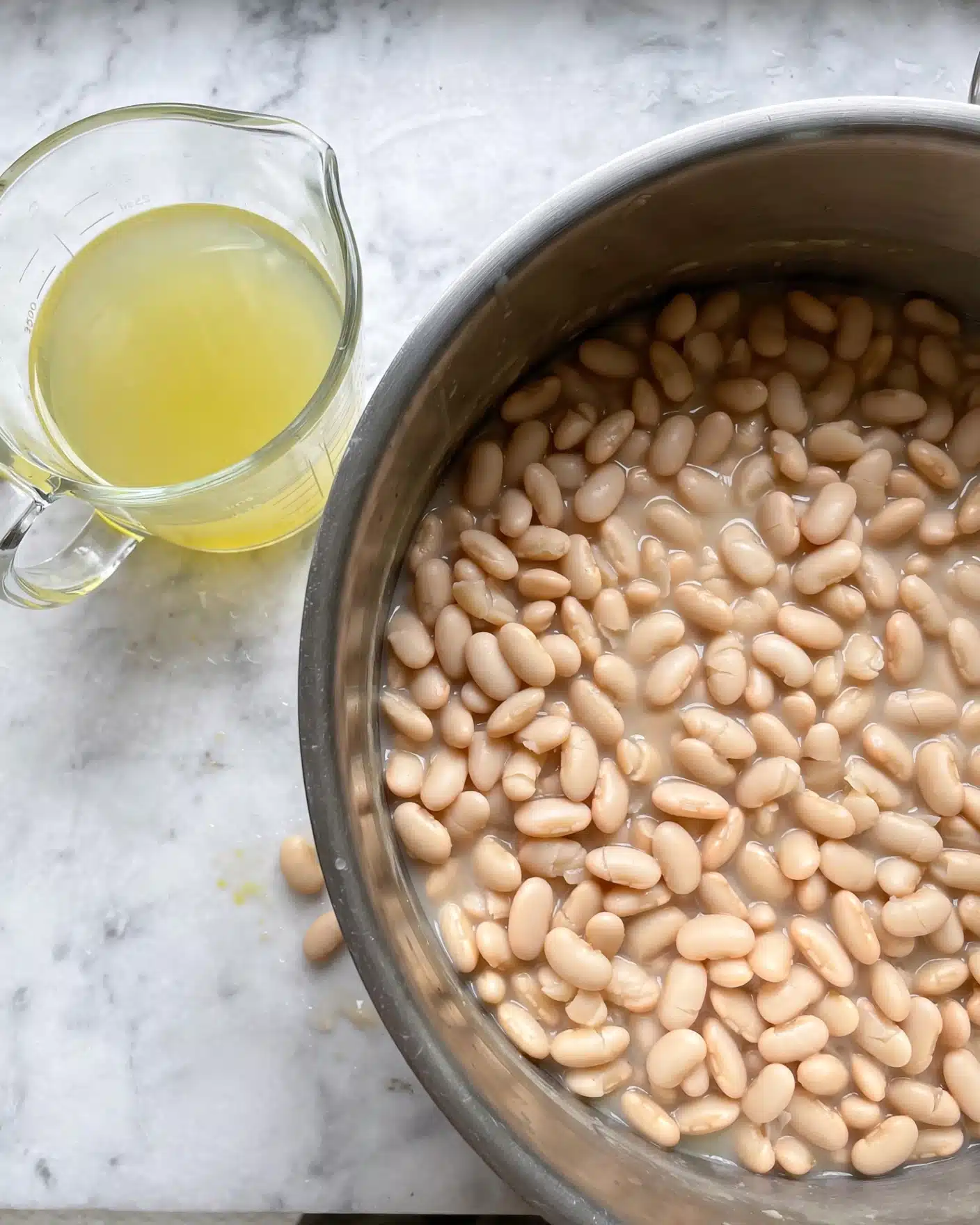 Grandma's Homemade Baked Beans Recipe 4 A large silver pot filled with soft white beans is placed on a white marbled surface, showing a close-up top view of the creamy textured beans filling the pot to the rim. Next to the pot on the left side is a clear glass measuring cup with a handle, containing pale yellowish liquid, sitting directly on the white marbled surface. The whole setup is simple and clean, highlighting the natural colors and textures of the beans and liquid. photo taken with an iphone --ar 4:5 --v 7