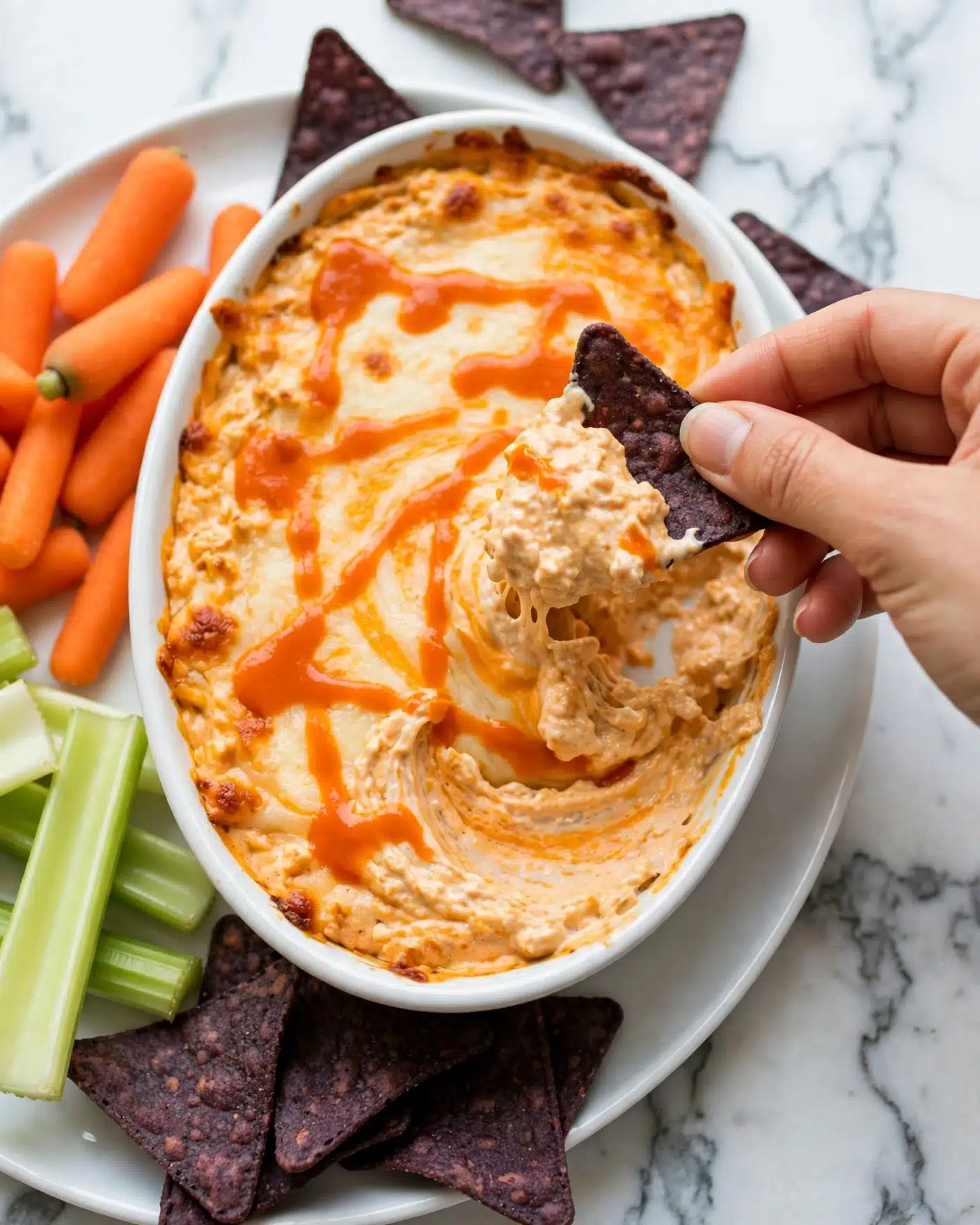 Healthy Buffalo Chicken Dip Recipe 5 A white oval dish filled with a creamy orange and white layered dip with a smooth melted cheese texture on top, drizzled with bright orange sauce in irregular lines. A woman's hand is dipping a dark purple corn chip loaded with the cheesy dip. Around the dish on a white plate there are fresh orange baby carrots, pale green celery sticks, and more dark purple corn chips. The background is a white marbled surface. Photo taken with an iphone --ar 4:5 --v 7