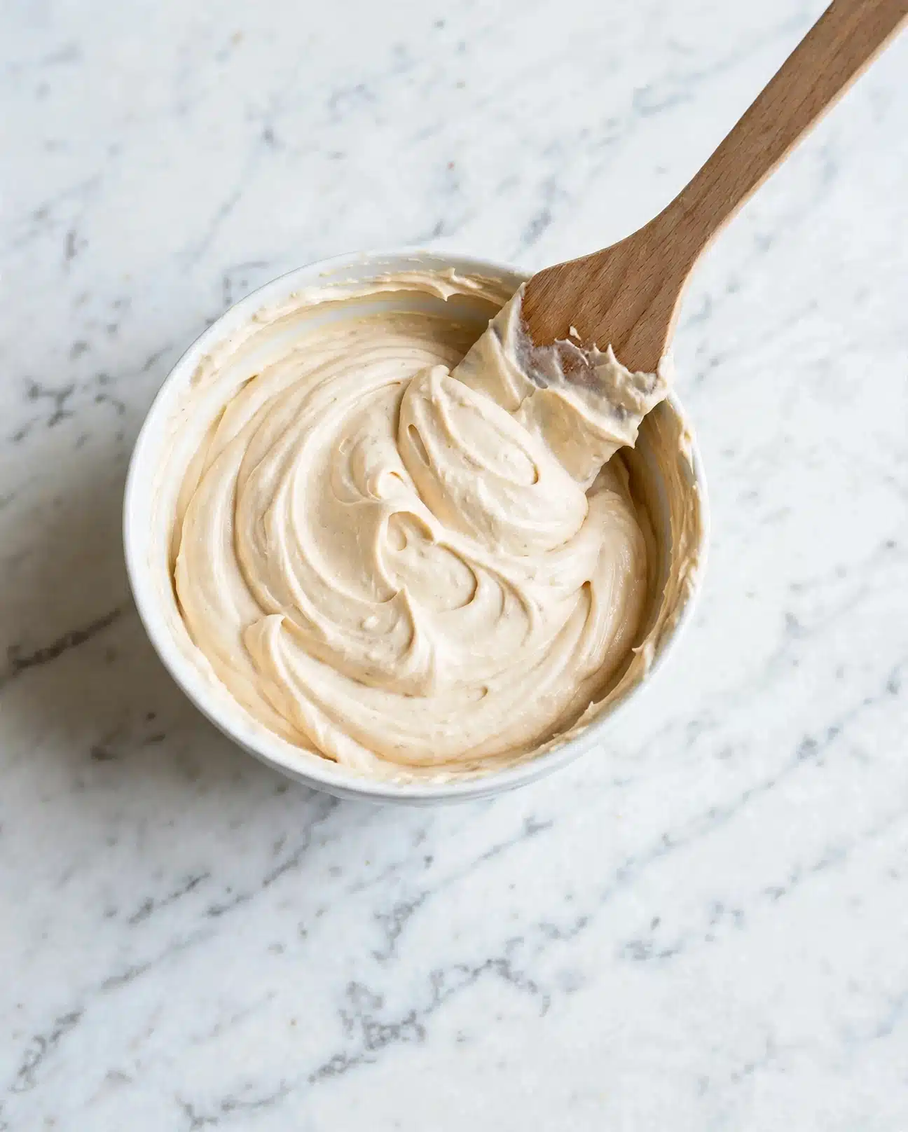 Dog Birthday Cake Recipe 4 A clear white bowl filled with smooth, light beige cream that is swirled and textured softly, showing thick and fluffy consistency. A wooden spatula is partially lifted from the bowl, with cream resting on its edge, surrounded by more cream spread evenly inside the bowl. The scene is set on a white marbled surface. photo taken with an iphone --ar 4:5 --v 7